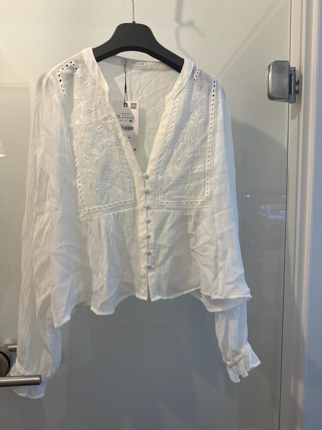White long-sleeved blouse with a V-neck, buttons and embroidery, hanging on a black hanger on a glass door.