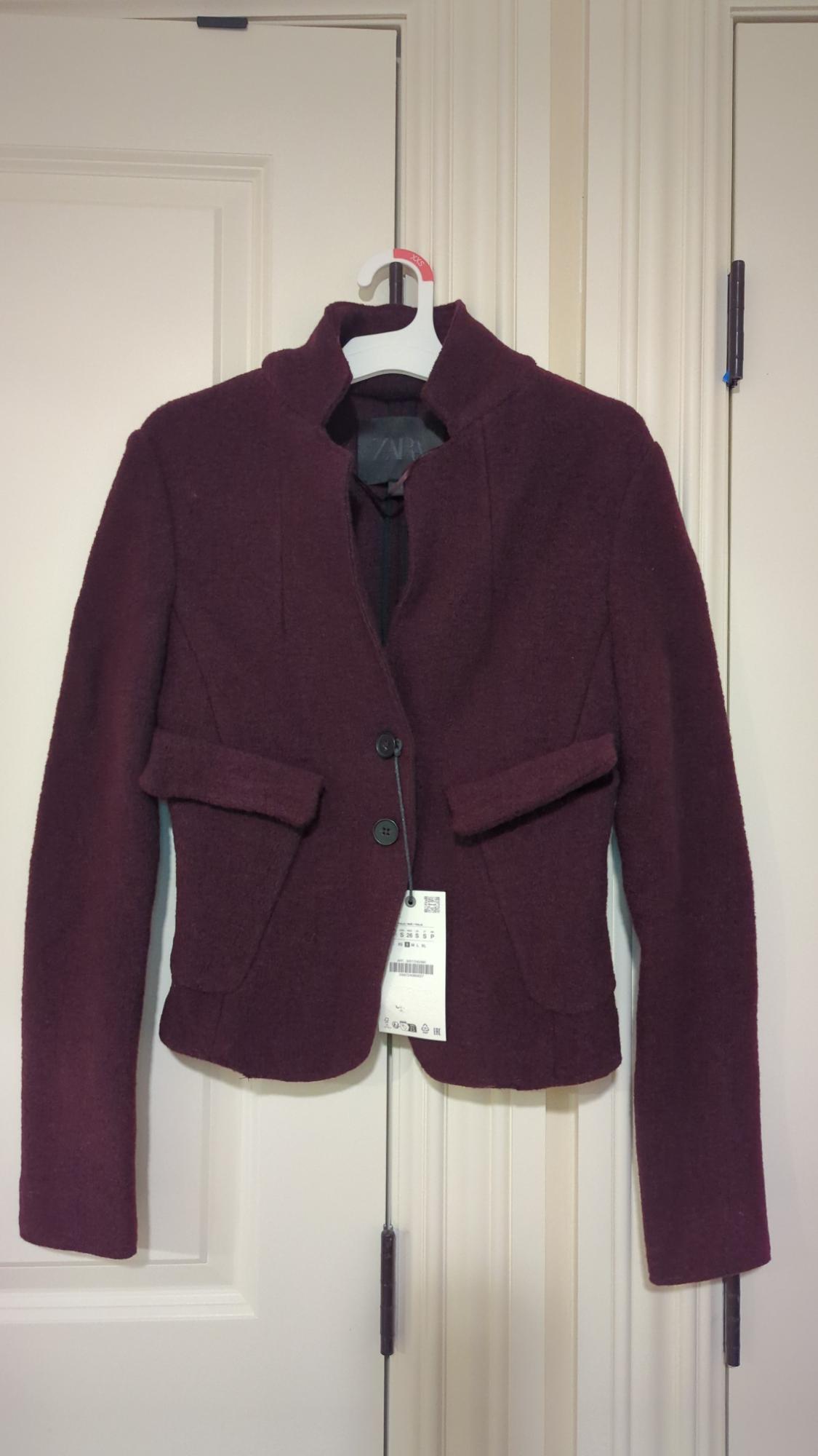 Burgundy wool jacket with two buttons and pockets, hanging on a white hanger over a white door. Label visible.