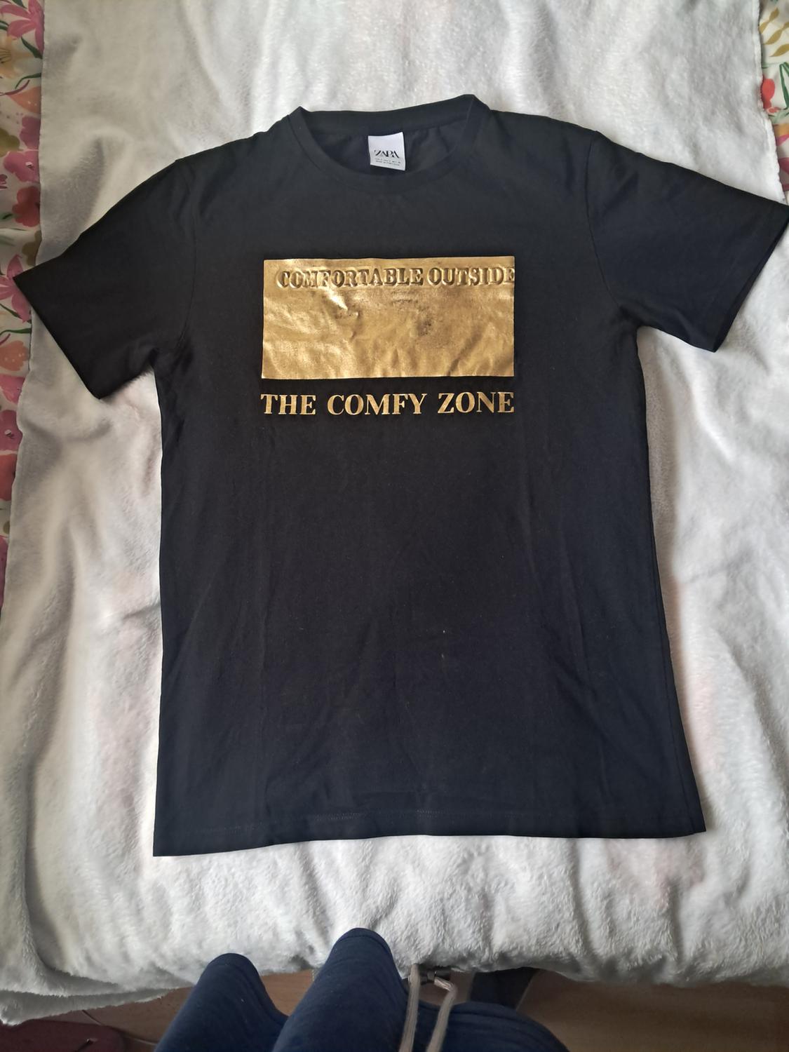Black T-shirt on a white blanket with a gold border and the text COMFORTABLE OUTSIDE THE COMFY ZONE.