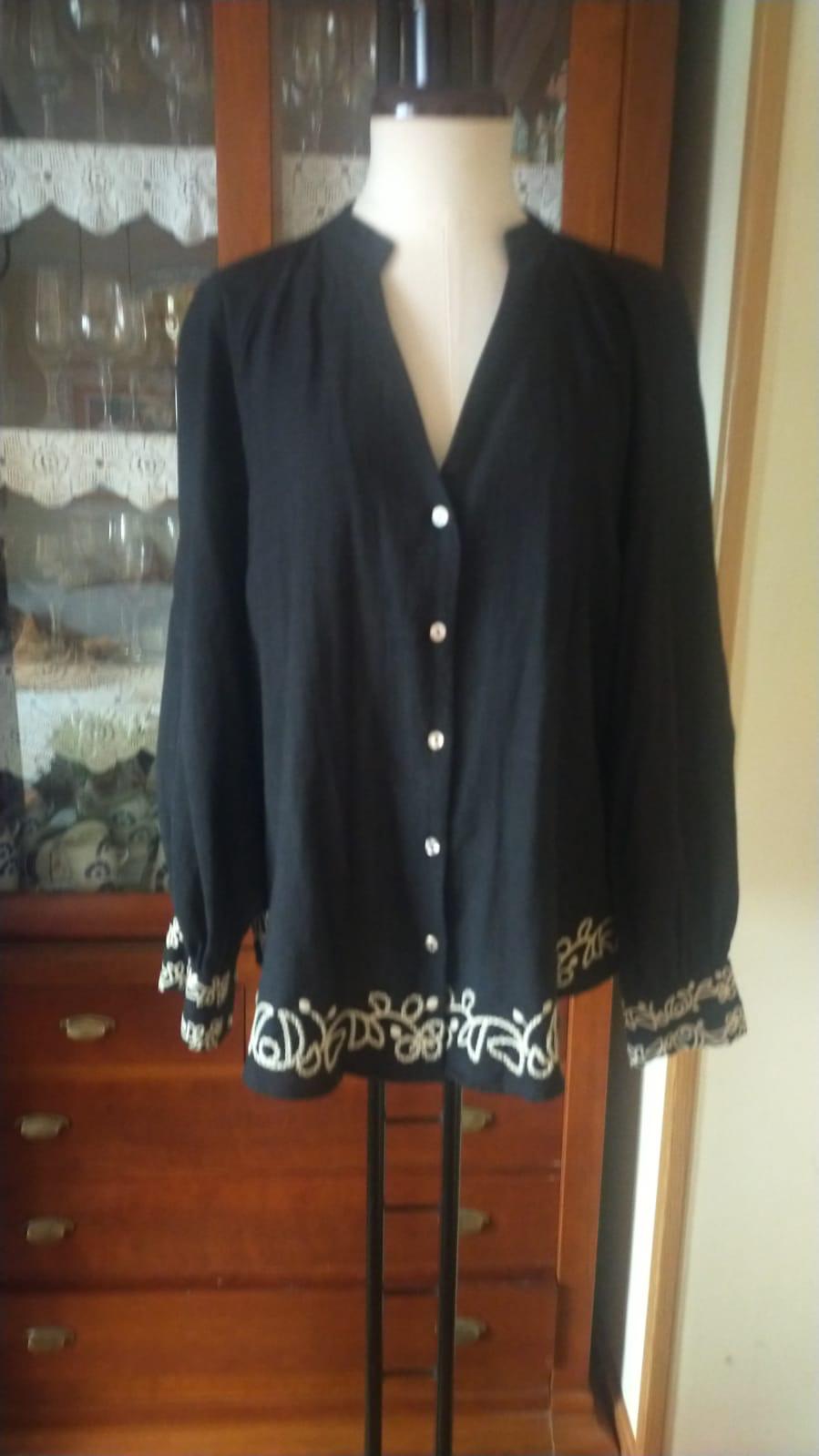 Black blouse with white buttons and white embroidery on the hem and cuffs, displayed on a mannequin in front of a wooden cabinet.