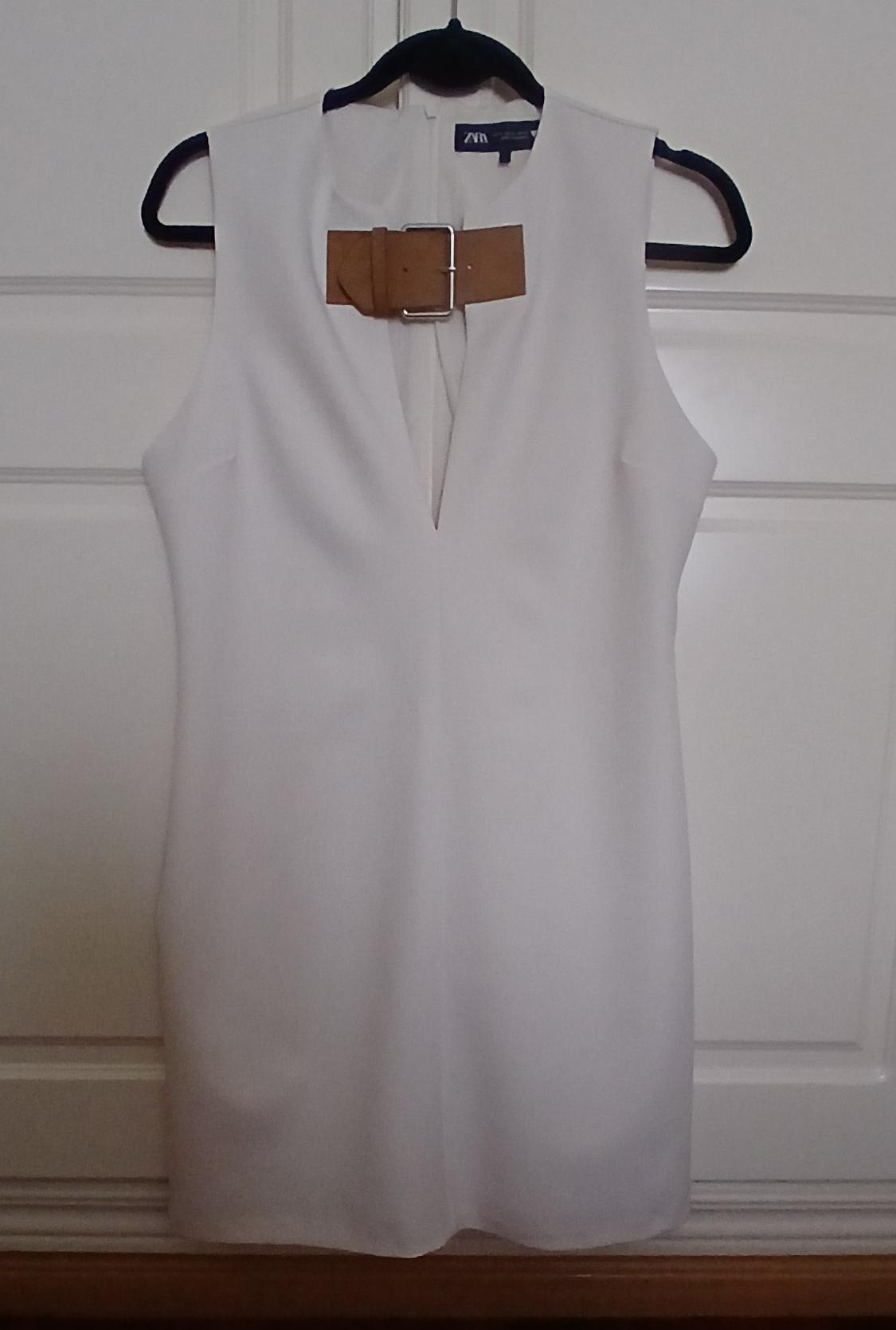 Sleeveless white dress hanging on a black hanger, with a brown belt and metal buckle at the neck. White door background.