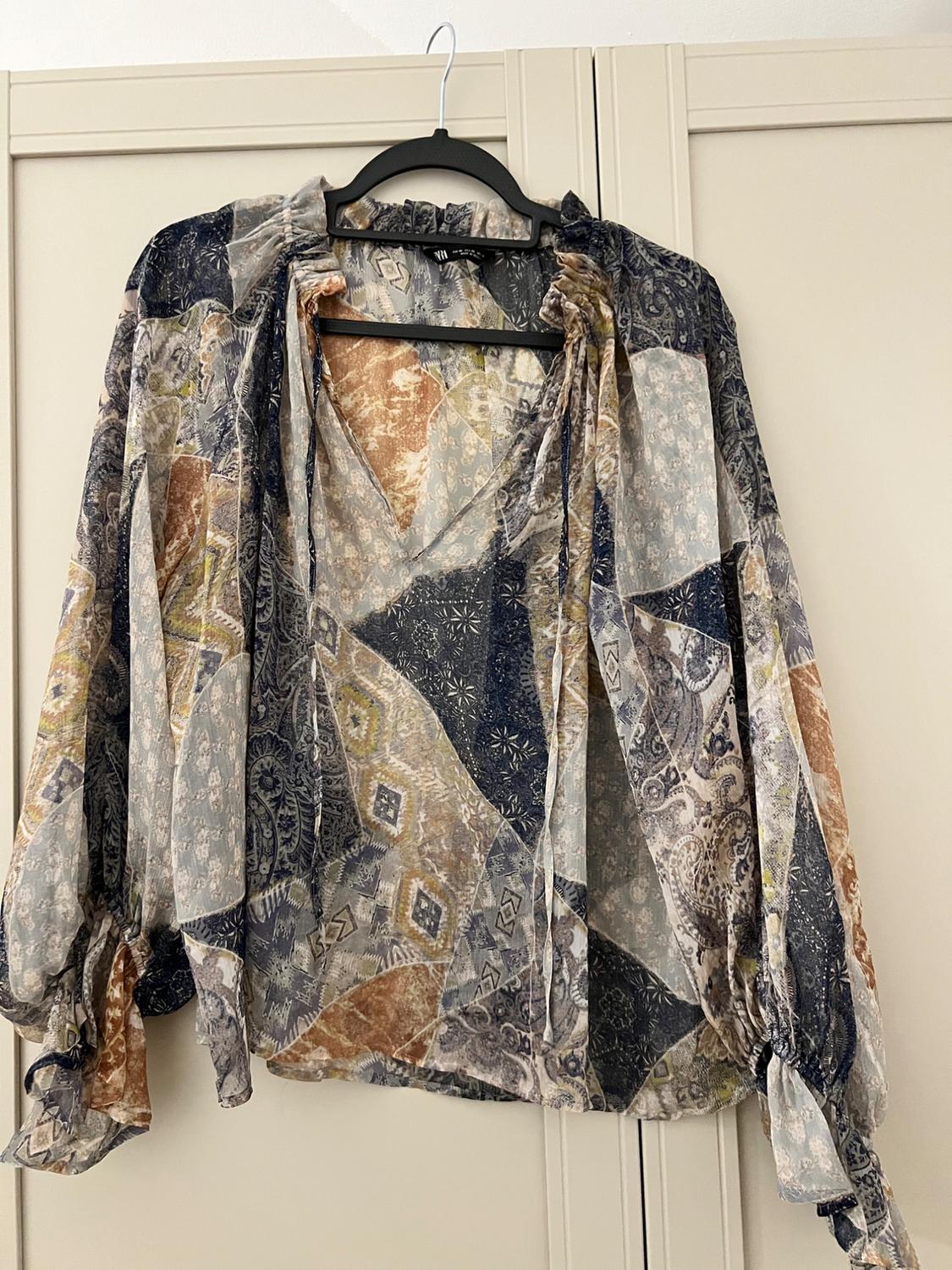 Chiffon blouse with a geometric print in shades of blue, brown, and beige, hanging on a black hanger in front of a light-coloured wardrobe.