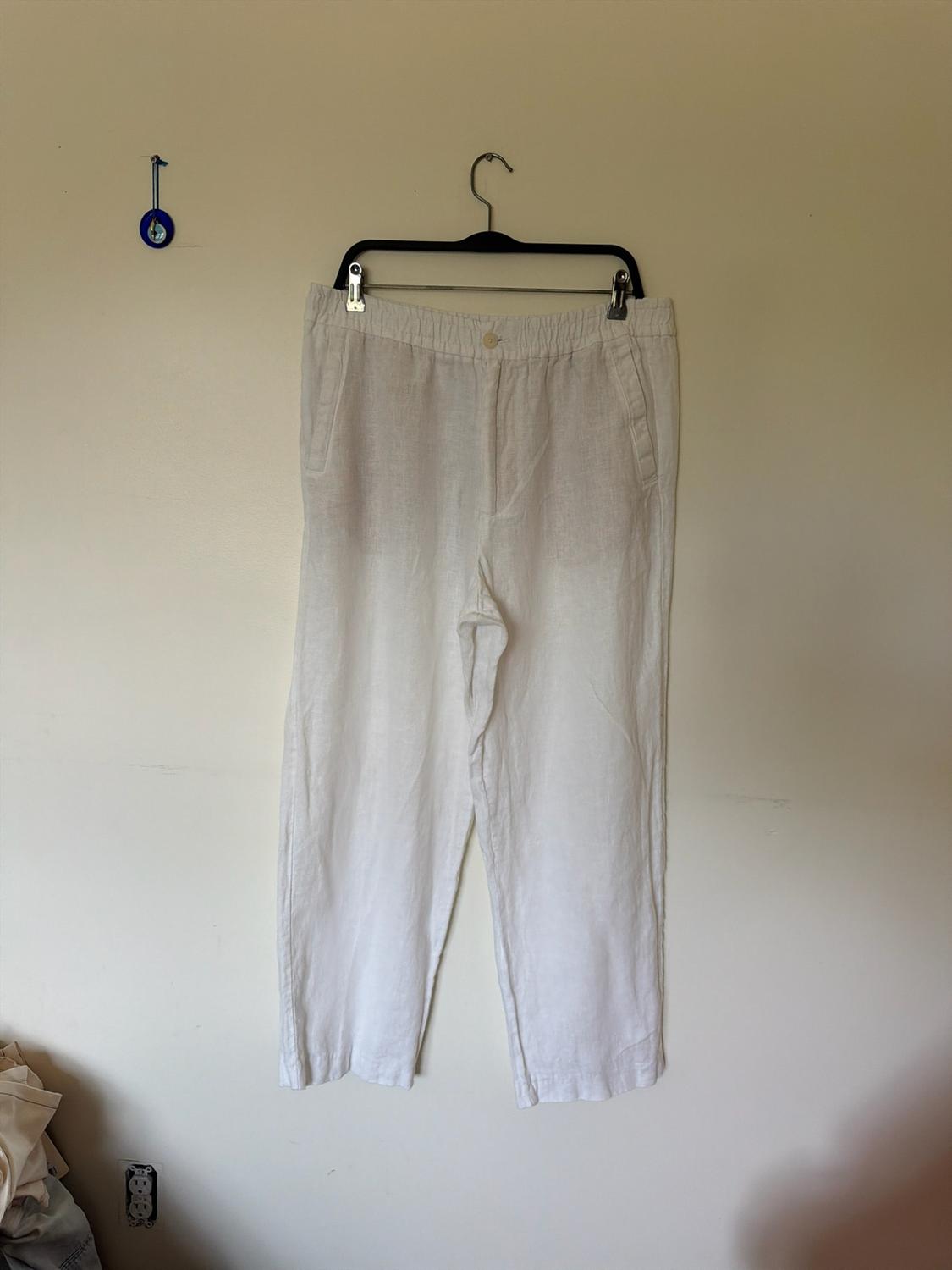 Loose white trousers hanging on a hanger against a beige wall. On the left, there is a small blue amulet and a plug socket at the bottom.