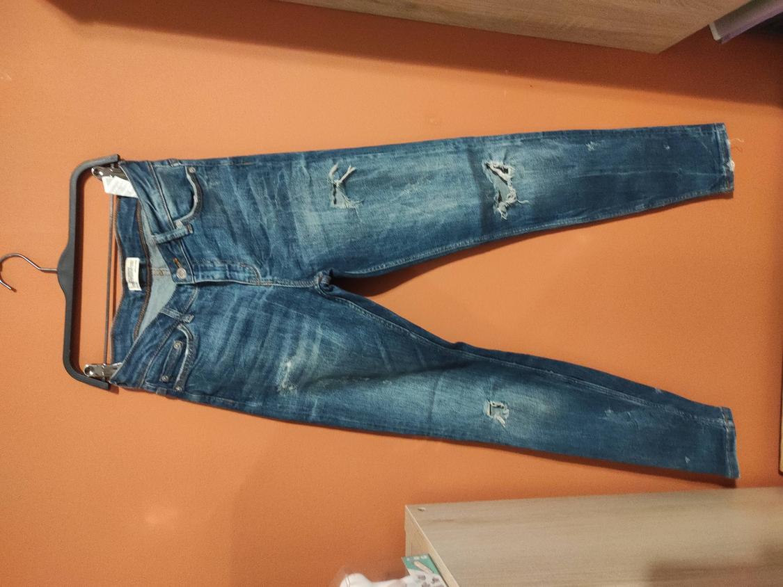 Worn blue jeans hanging on a black hanger against an orange wall. Ripped details on the legs.
