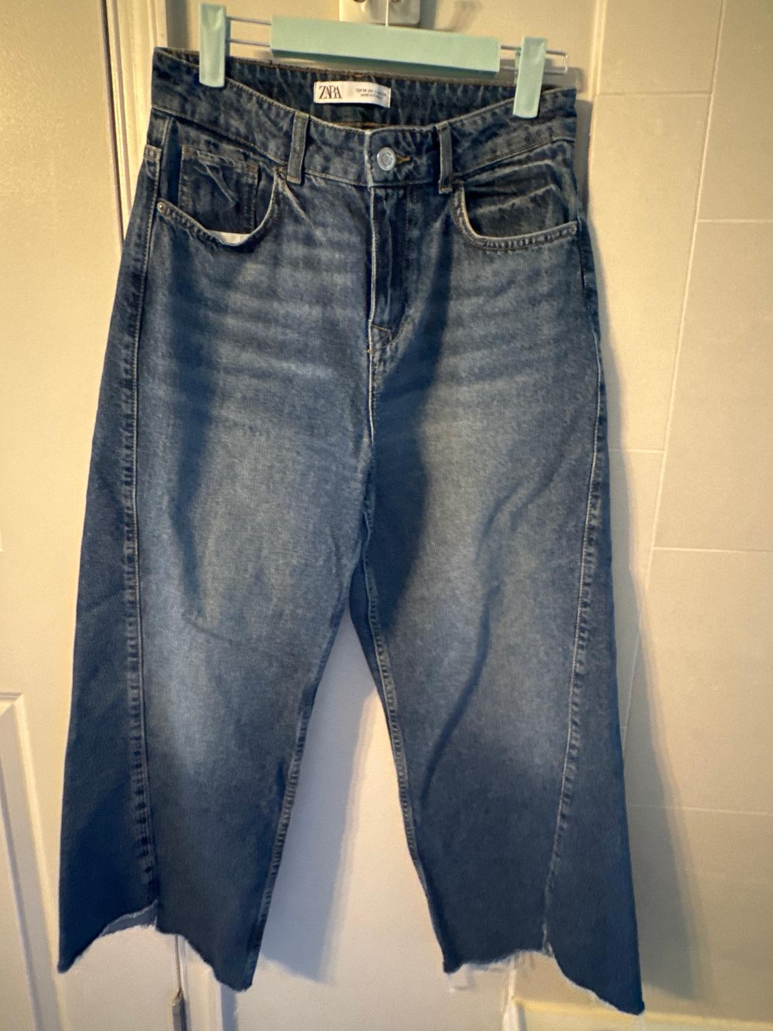 Blue straight-cut jeans hanging on a green hanger. Visible label on the waistband with the text "ZARA". White wall background.