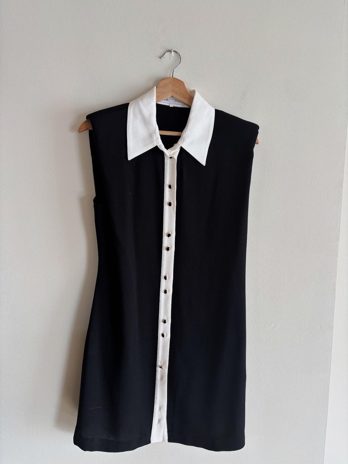 Sleeveless black dress with a collar and white buttons, hanging on a wooden hanger against a white wall.