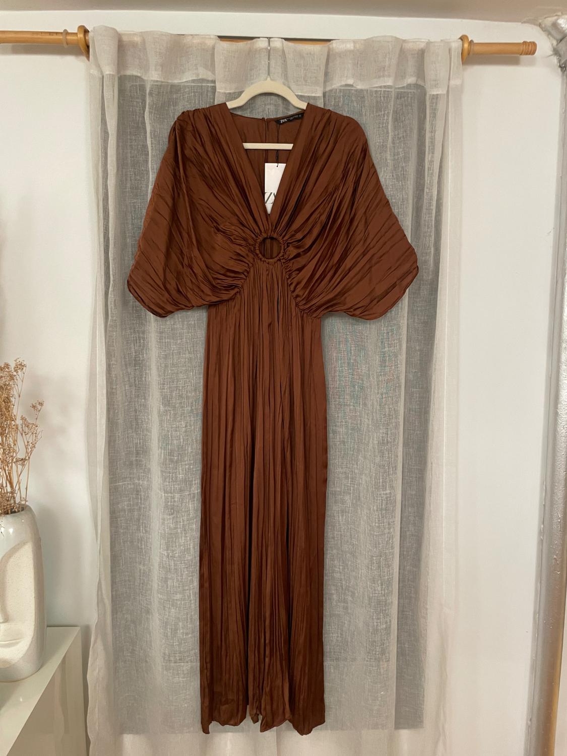 Pleated brown dress with wide sleeves and a ring detail on the chest, hanging on a hanger in front of a white curtain.