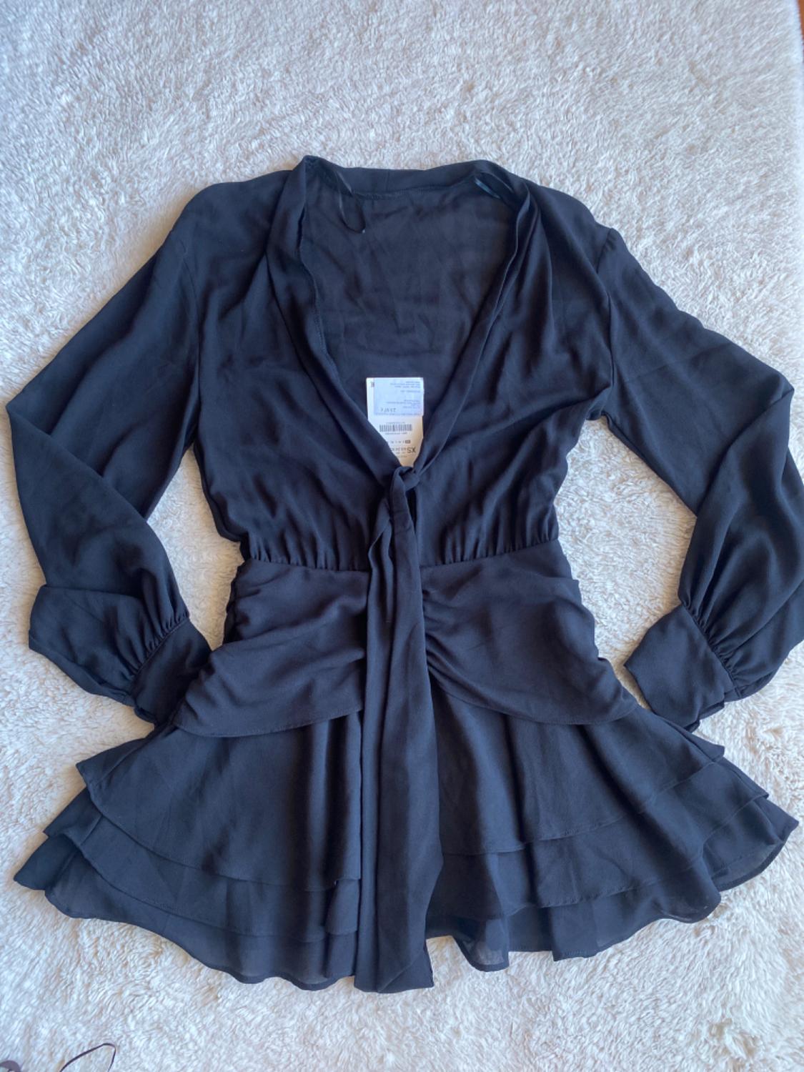 Black long-sleeved blouse with a tie waist and frills on the hem, on a light surface. Visible label in the centre.