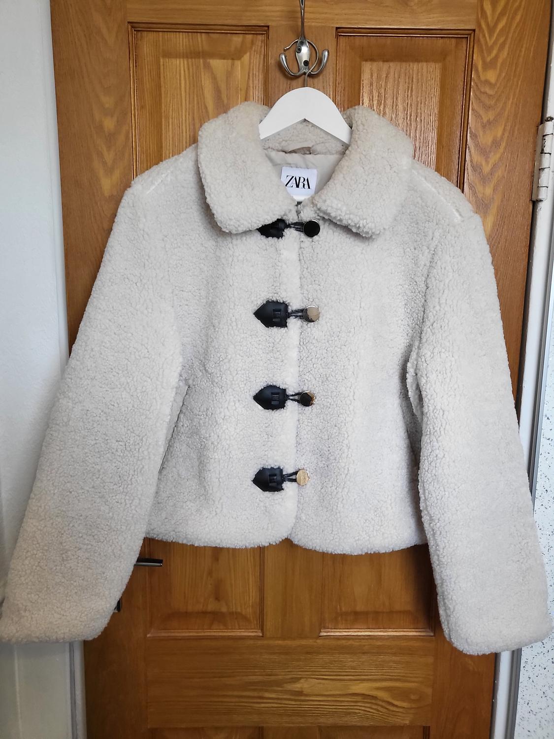 Beige wool jacket with large black buttons hanging on a wooden door. Visible label with text "ZARA".