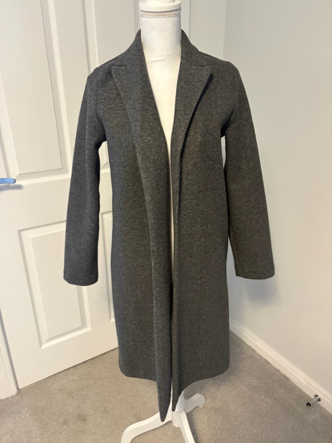 Dark grey long open coat with lapels on a white mannequin. In the background, a room with a white door and a grey rug.