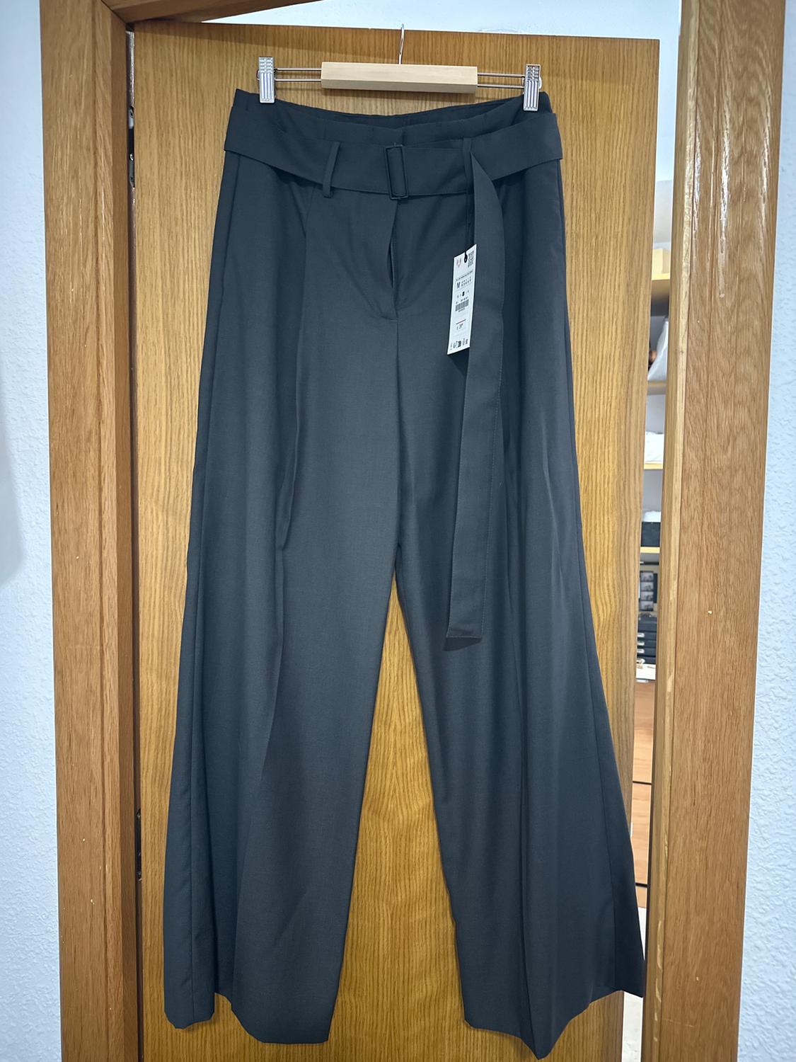 Grey wide-leg trousers hanging on a wooden door, with a matching belt and visible tag. Indoor room background.