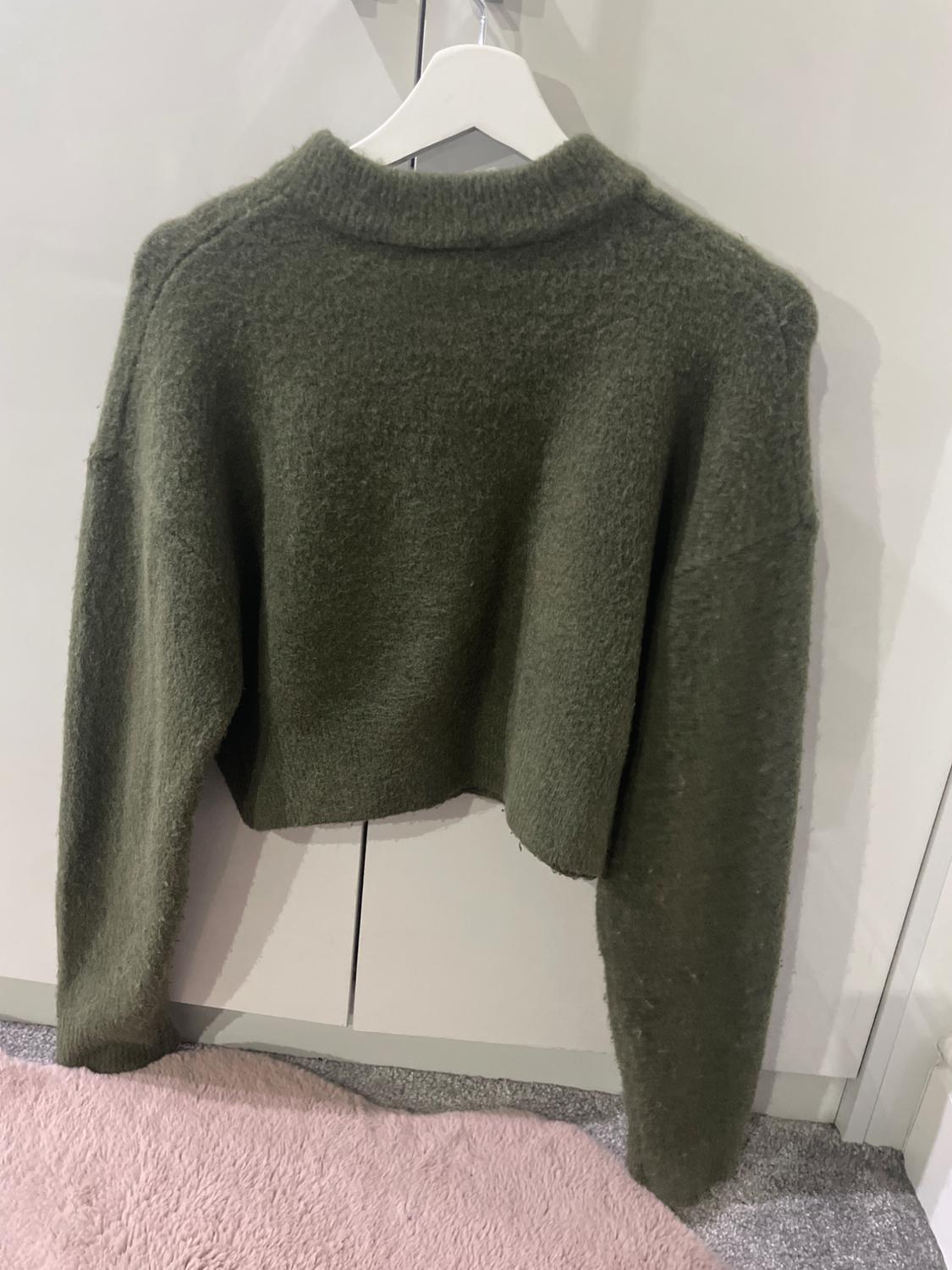 Olive green wool jumper hanging on a white hanger, against a white wardrobe background and pink rug.