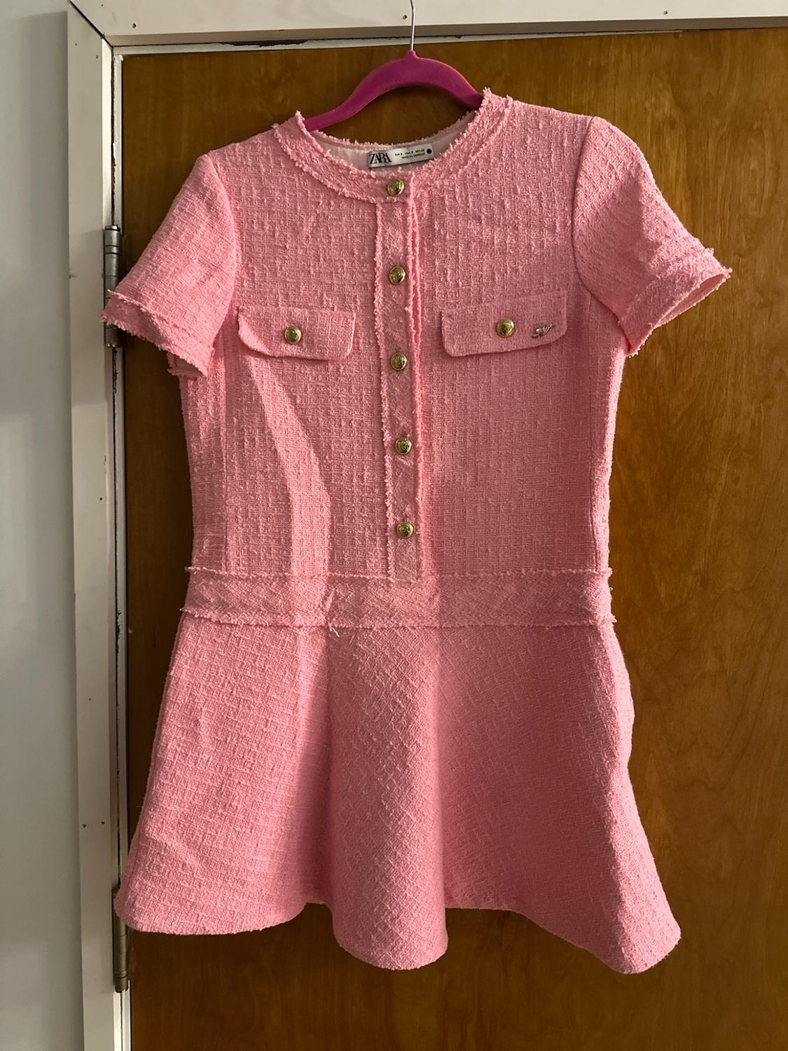 Pink short-sleeved textured dress with gold buttons and frayed hem, hanging on a wooden door. ZARA text.