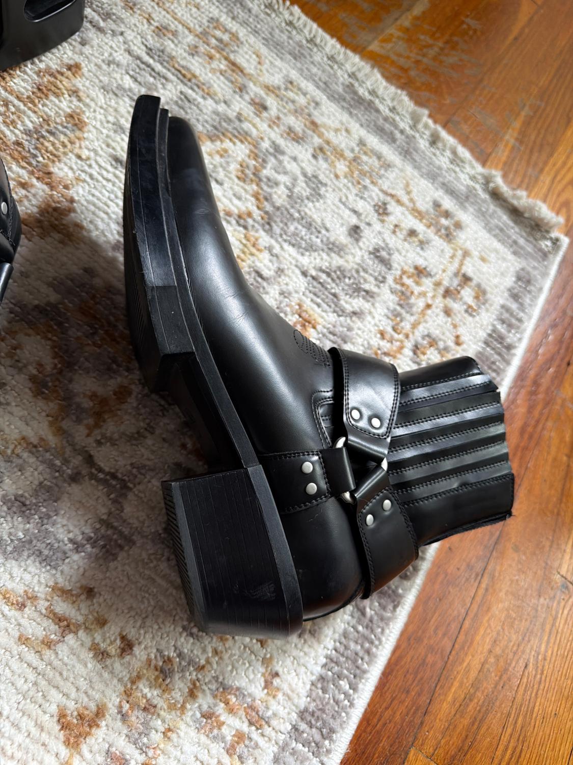 Black boot with straps, ring, and silver studs, resting on its side on a patterned rug and wooden floor.