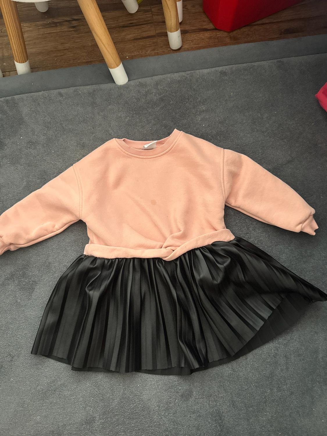 Dress on a gray rug. Long-sleeved pink sweatshirt with a knotted waist and a black pleated skirt. Wooden chair legs in the background.
