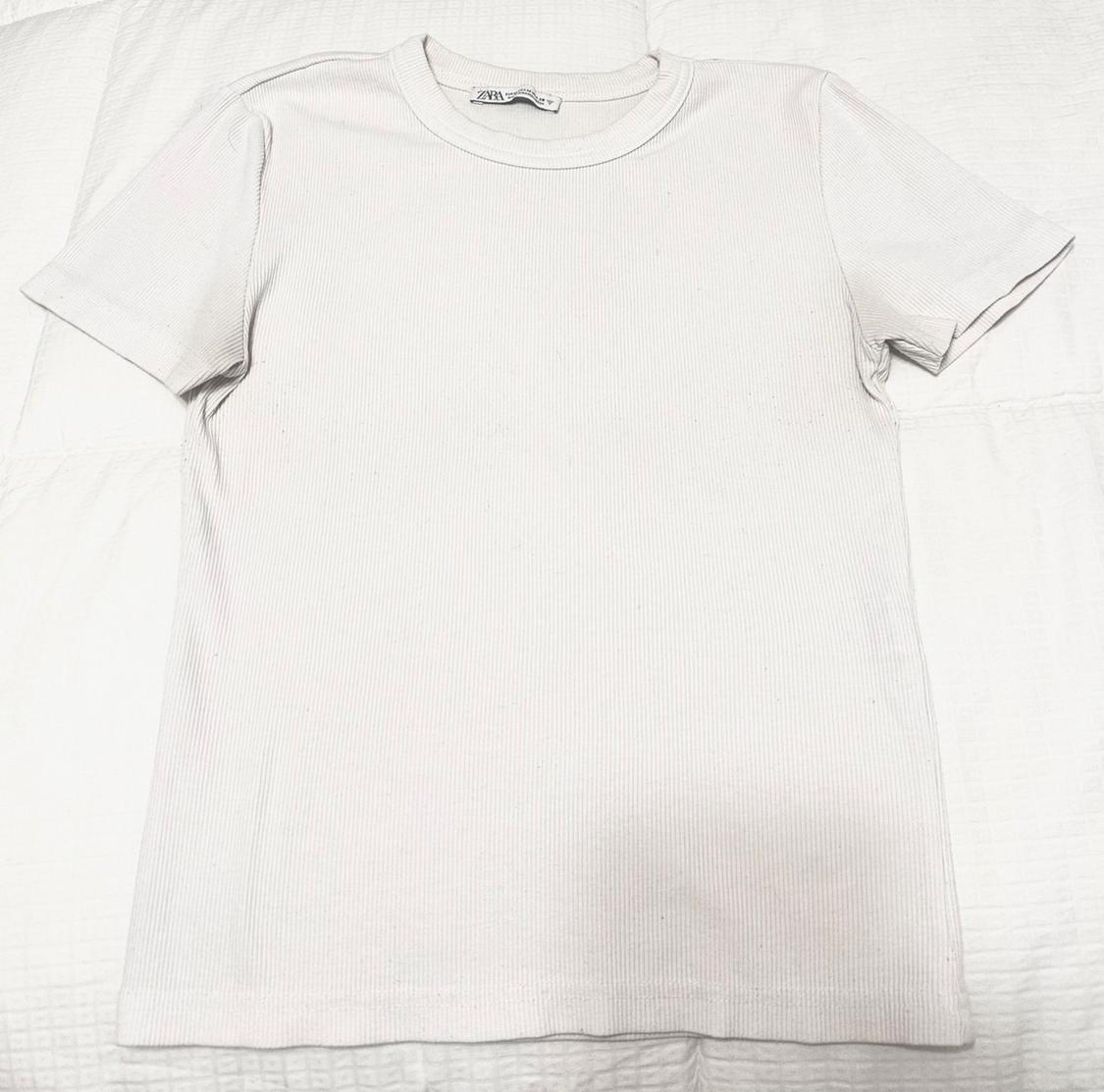 White short-sleeve T-shirt with a ribbed texture, laid out on a light-colored fabric surface. Label with the text ZARA visible.