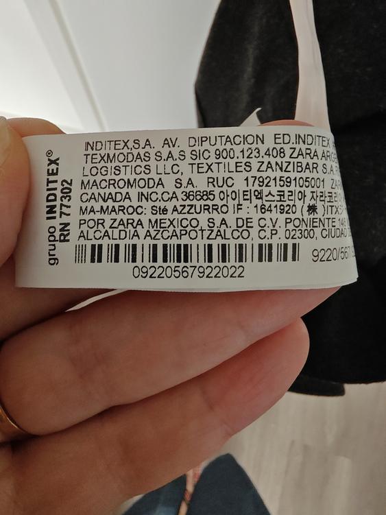 Clothing label with detailed manufacturing and distribution text, including "INDITEX", "ZARA", and barcodes. Light wood background.