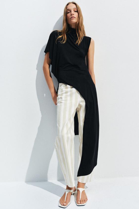 Souprava černé asymetrické halenky bez rukávů a bílých kalhot s vertikálními béžovými pruhy. Bílé sandály.