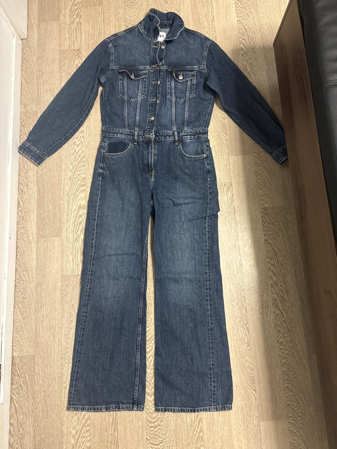 Blue long-sleeved denim jumpsuit with front buttons, chest pockets, and wide-leg trousers on a light wooden floor.