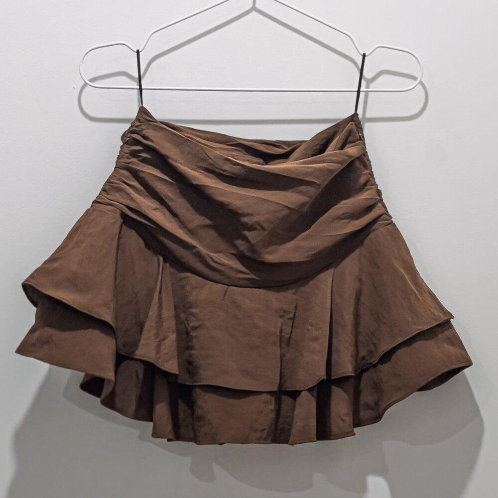 Short brown dress with ruffles at the hem and a cinched waist, hanging on a white hanger against a light-colored wall.