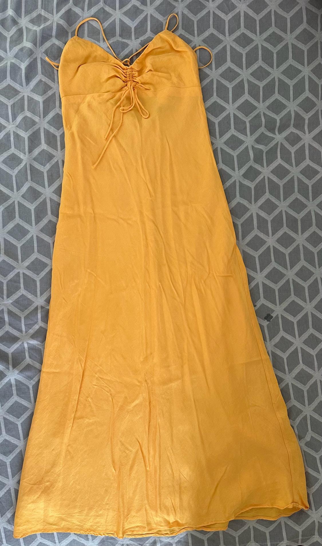 Long yellow thin-strapped dress with gathered detailing and ties on the neckline, laid out on a grey surface with a geometric pattern.
