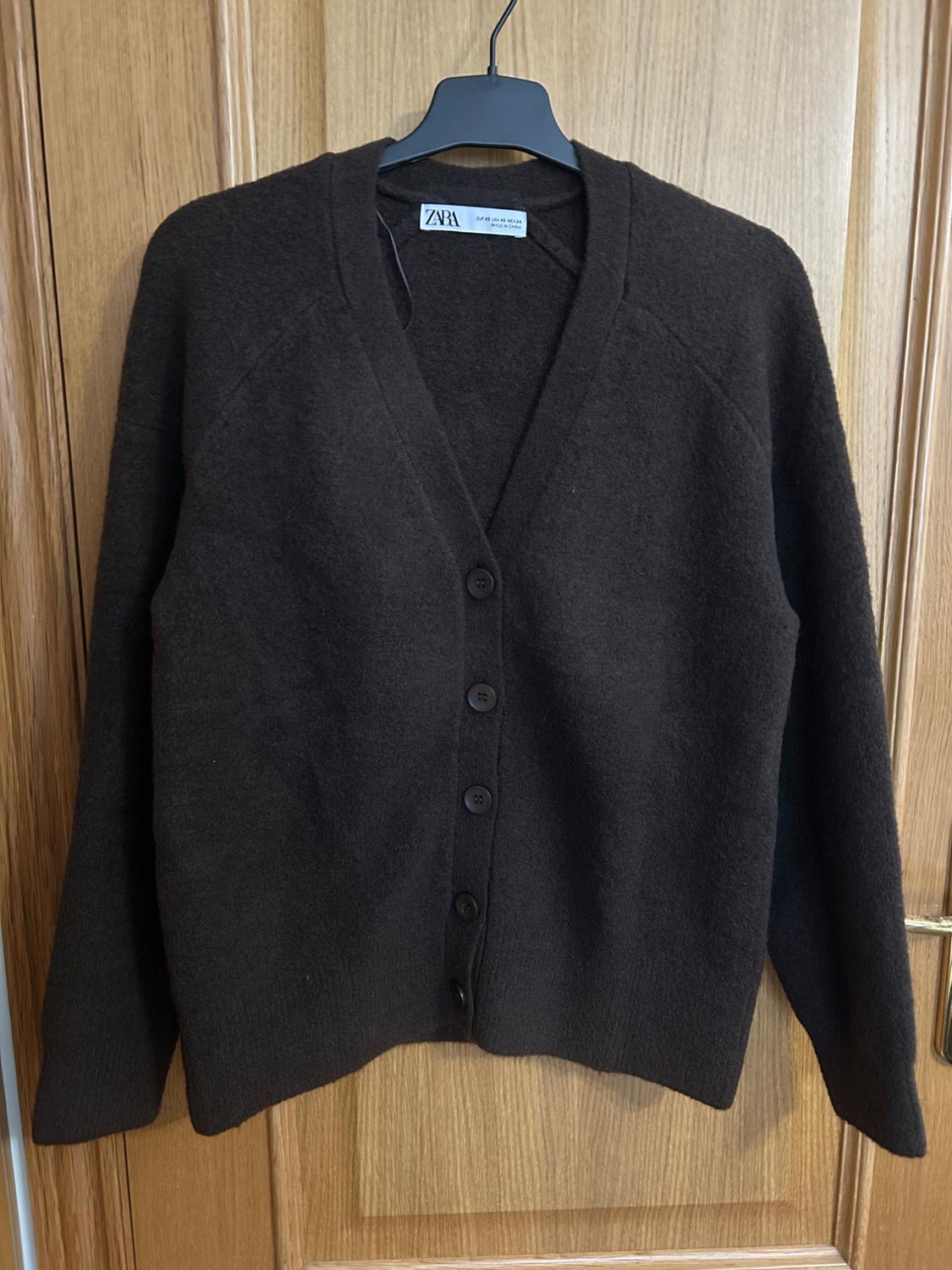 Black wool cardigan with buttons, hanging on a black hanger in front of a wooden door. Visible label: "ZARA".