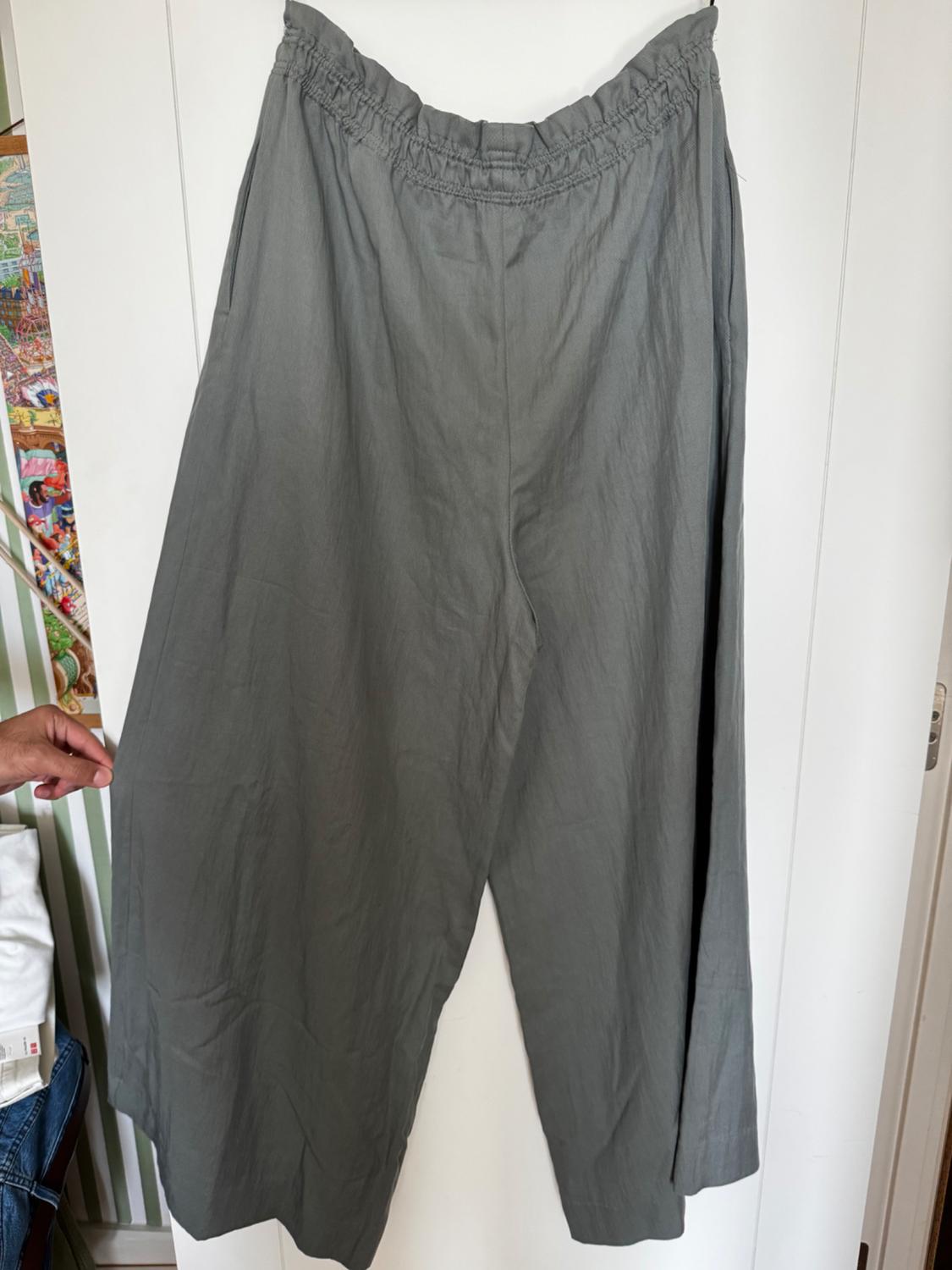Grey wide-leg trousers hang from a white door. A hand is holding the left side. The background has green and white stripes.