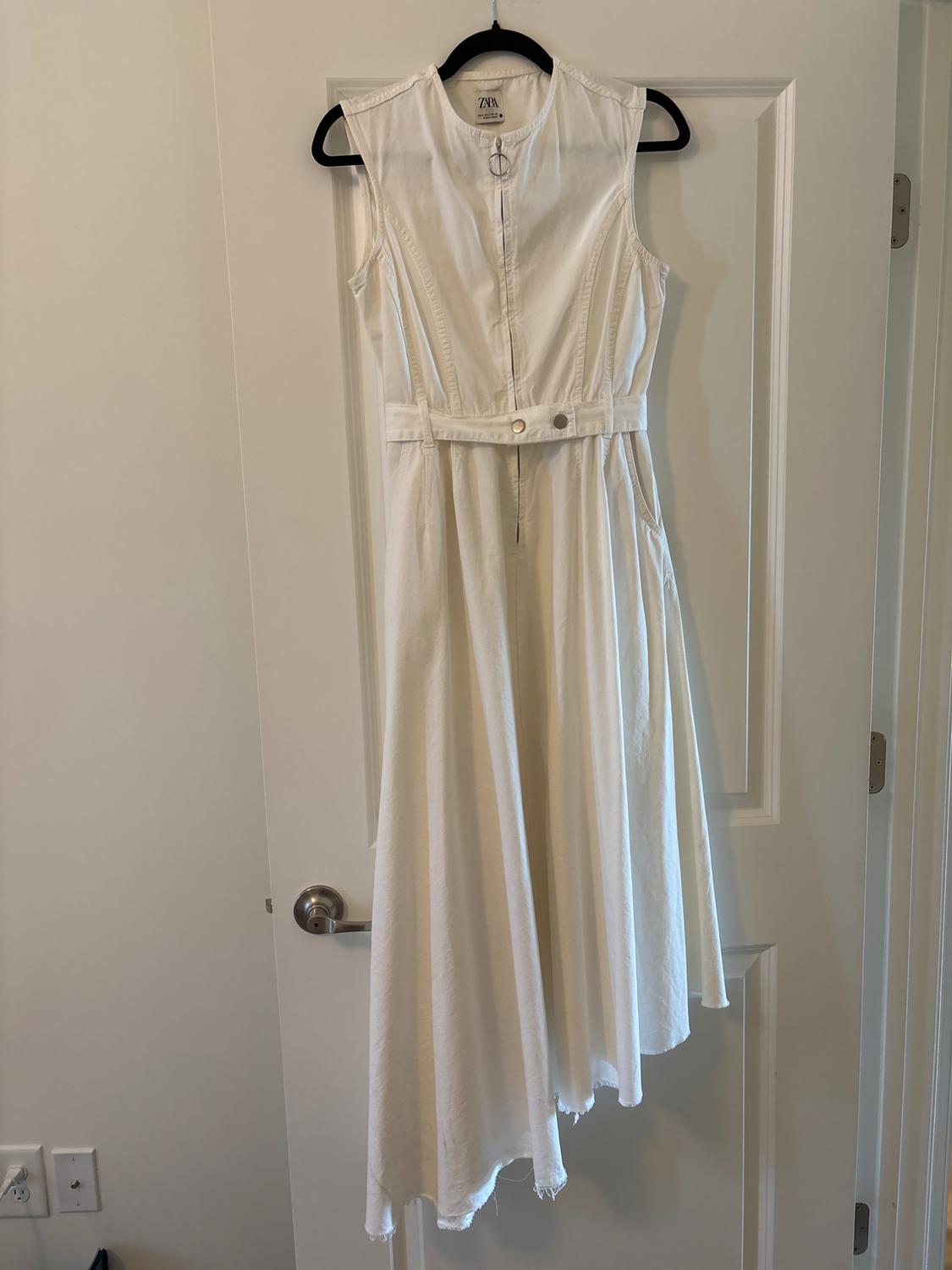 Sleeveless white dress hanging on a black hanger over a white door. It has a belt and front fastening.