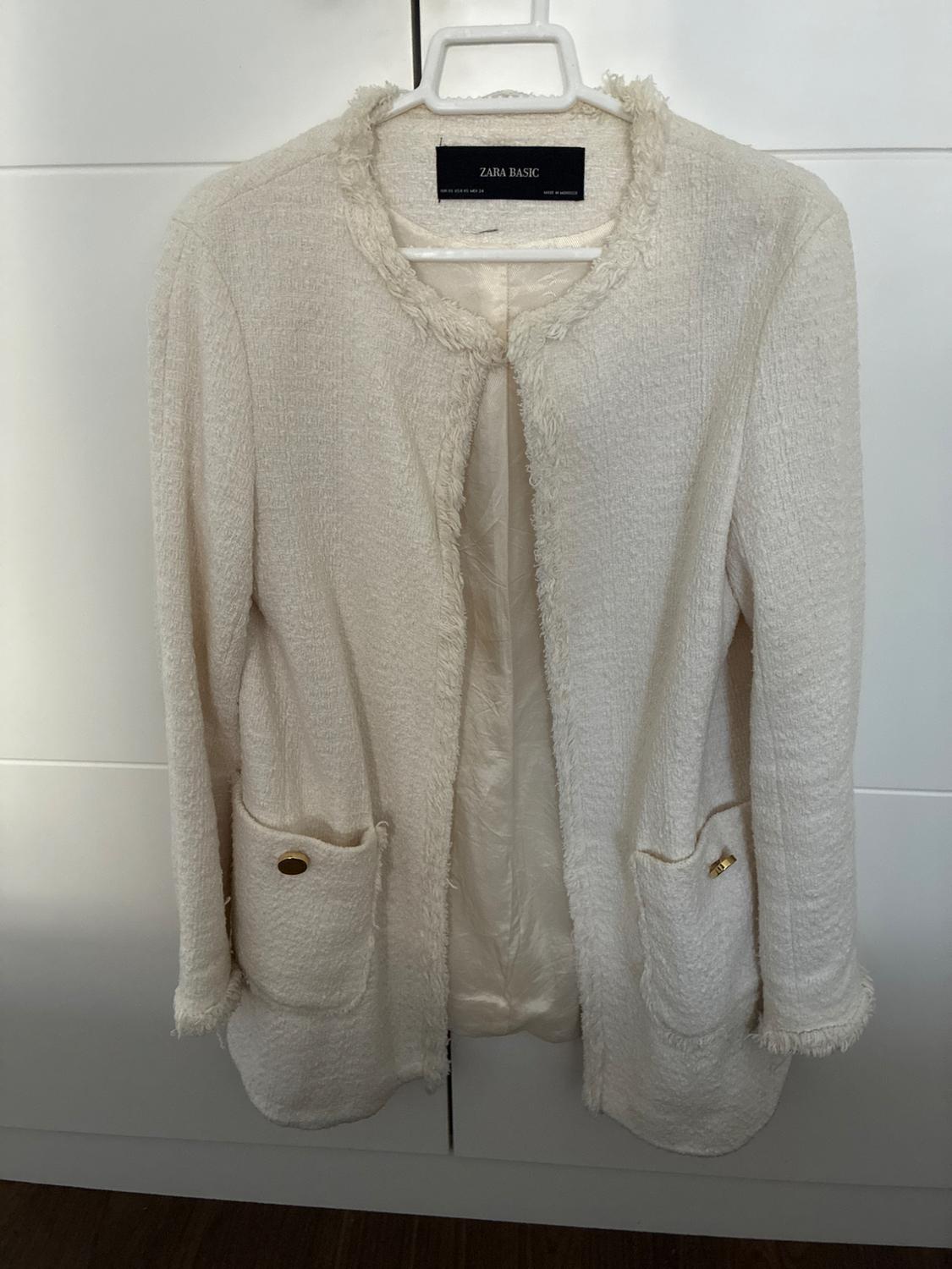 White textured jacket with frayed edges and gold button pockets, hanging on a hanger. ZARA BASIC label visible.