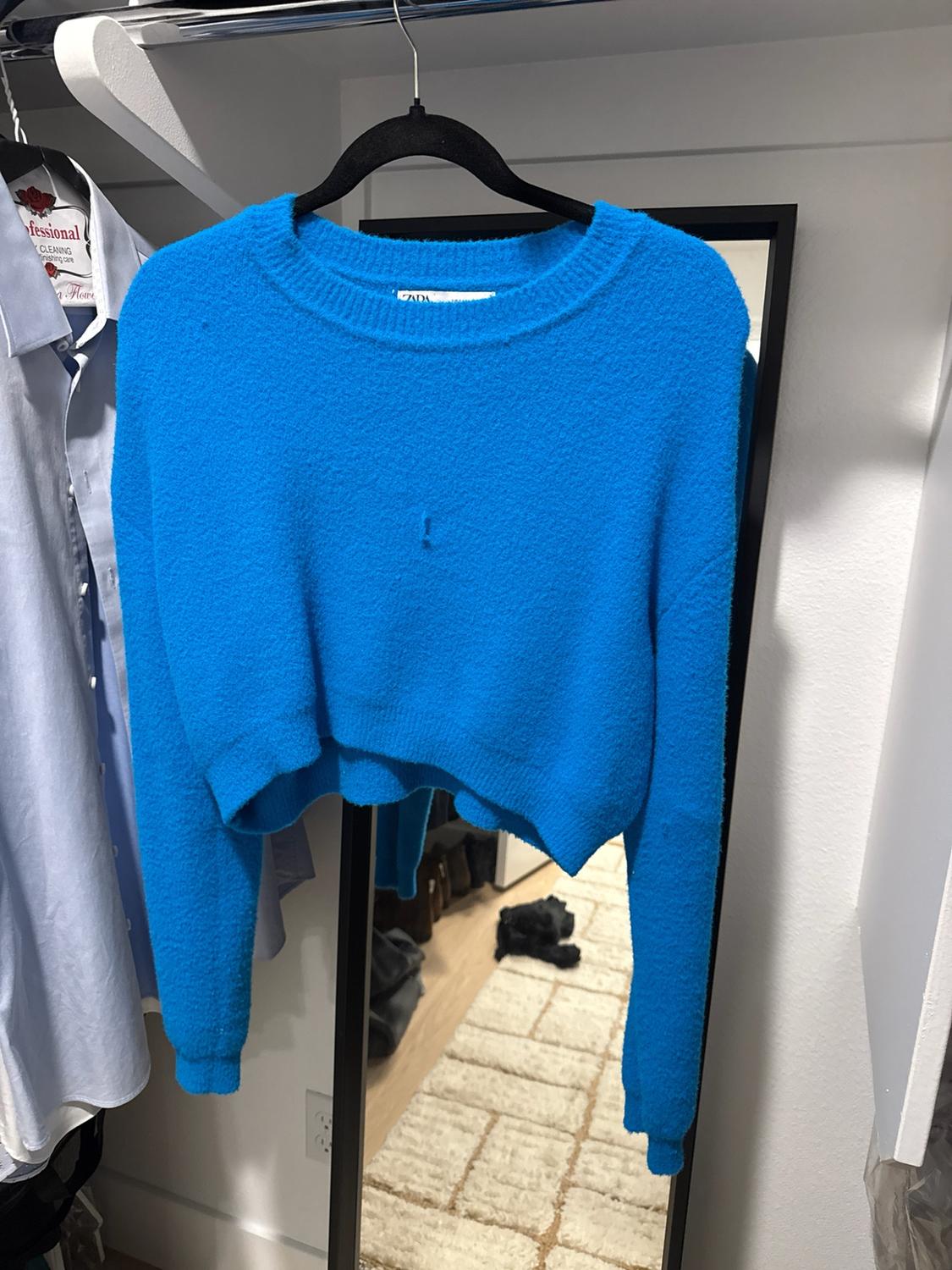 A blue knit sweater hanging in a closet in front of a mirror. Text: ZARA, fessional CLEANING nishing care.