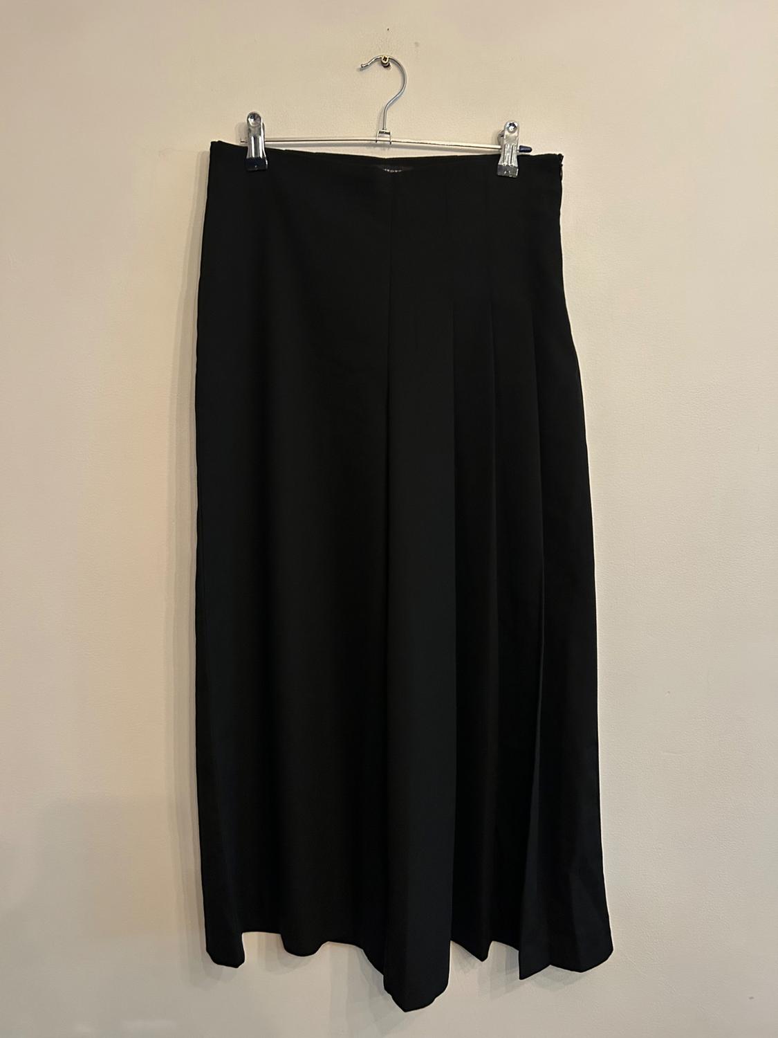 Long black trousers hanging on a hanger against a beige wall. The design is simple and elegant, with subtle pleats.