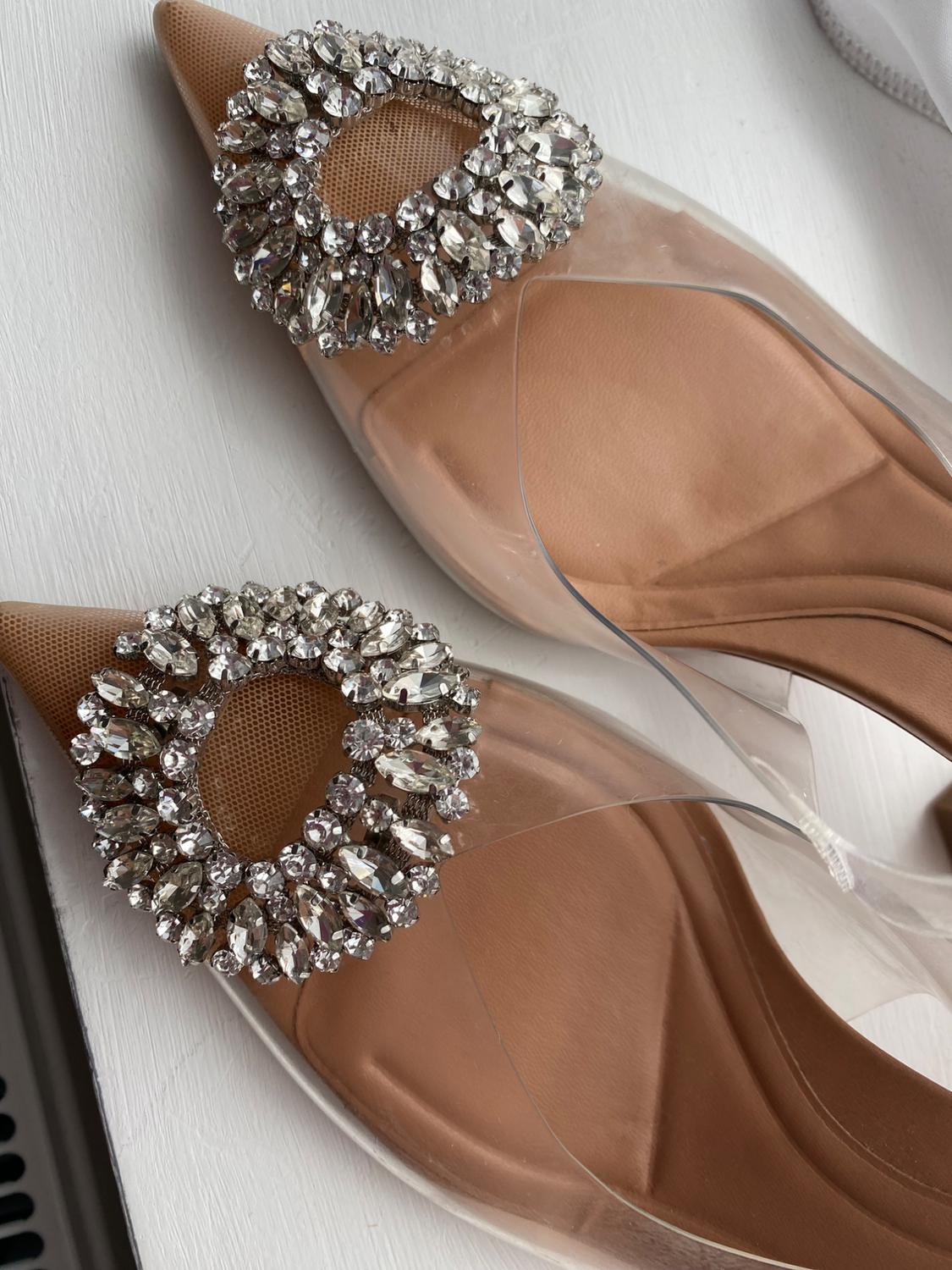 Close-up of pointed-toe shoes with transparent instep and circular embellishment of shiny rhinestones, on a white surface.