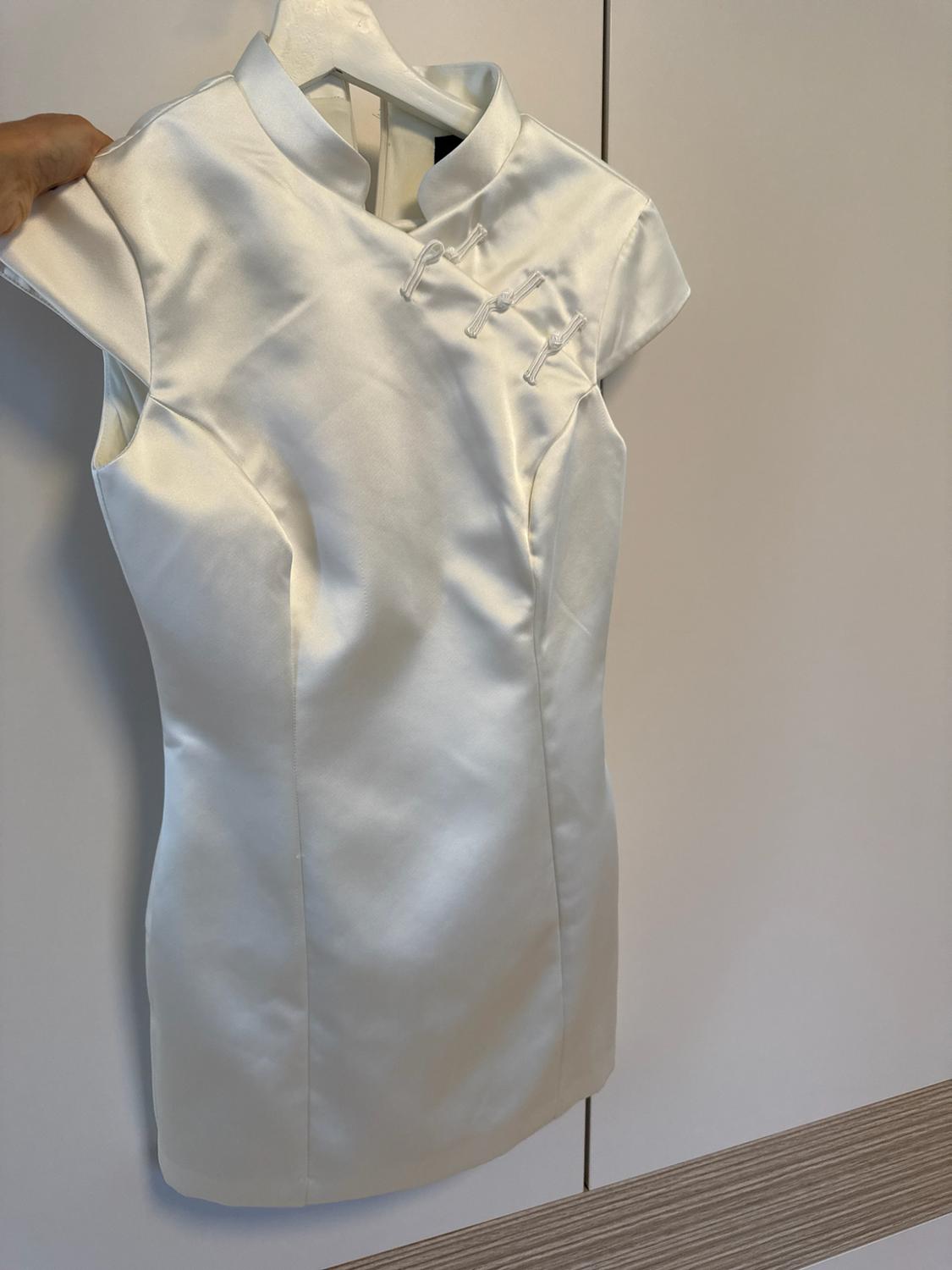 Hand holding a short white satin hanger dress with short sleeves, a mandarin collar and knot buttons, against a light background.