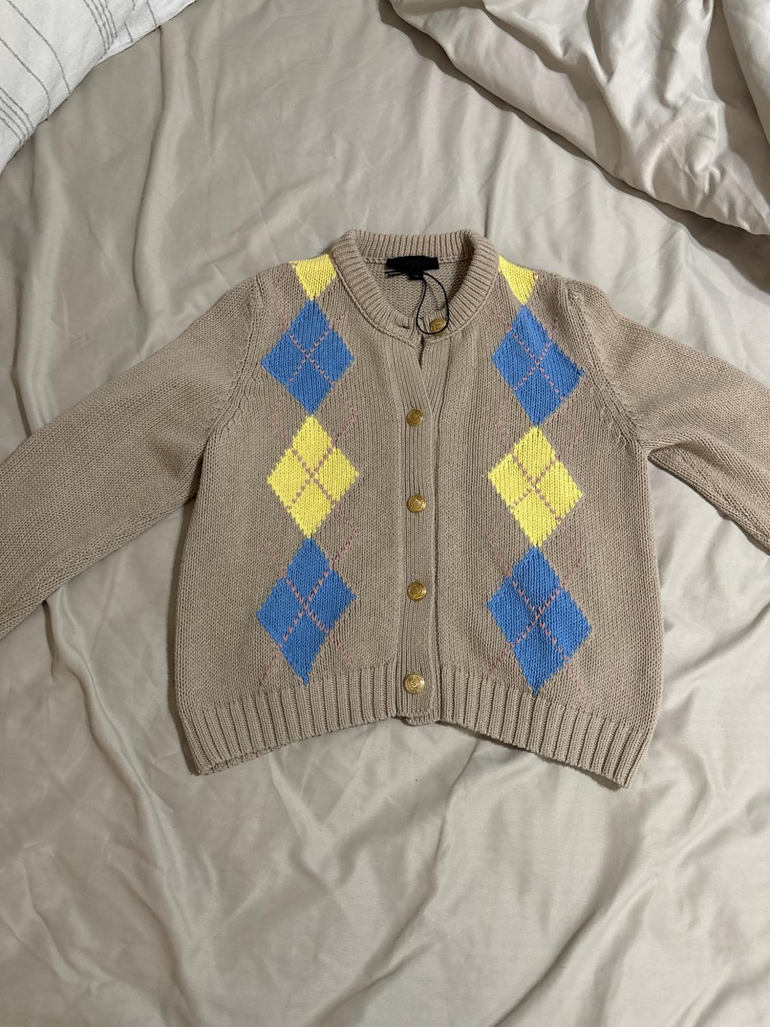Beige knitted cardigan with blue and yellow diamonds and gold buttons, spread out on some crumpled pale sheets.