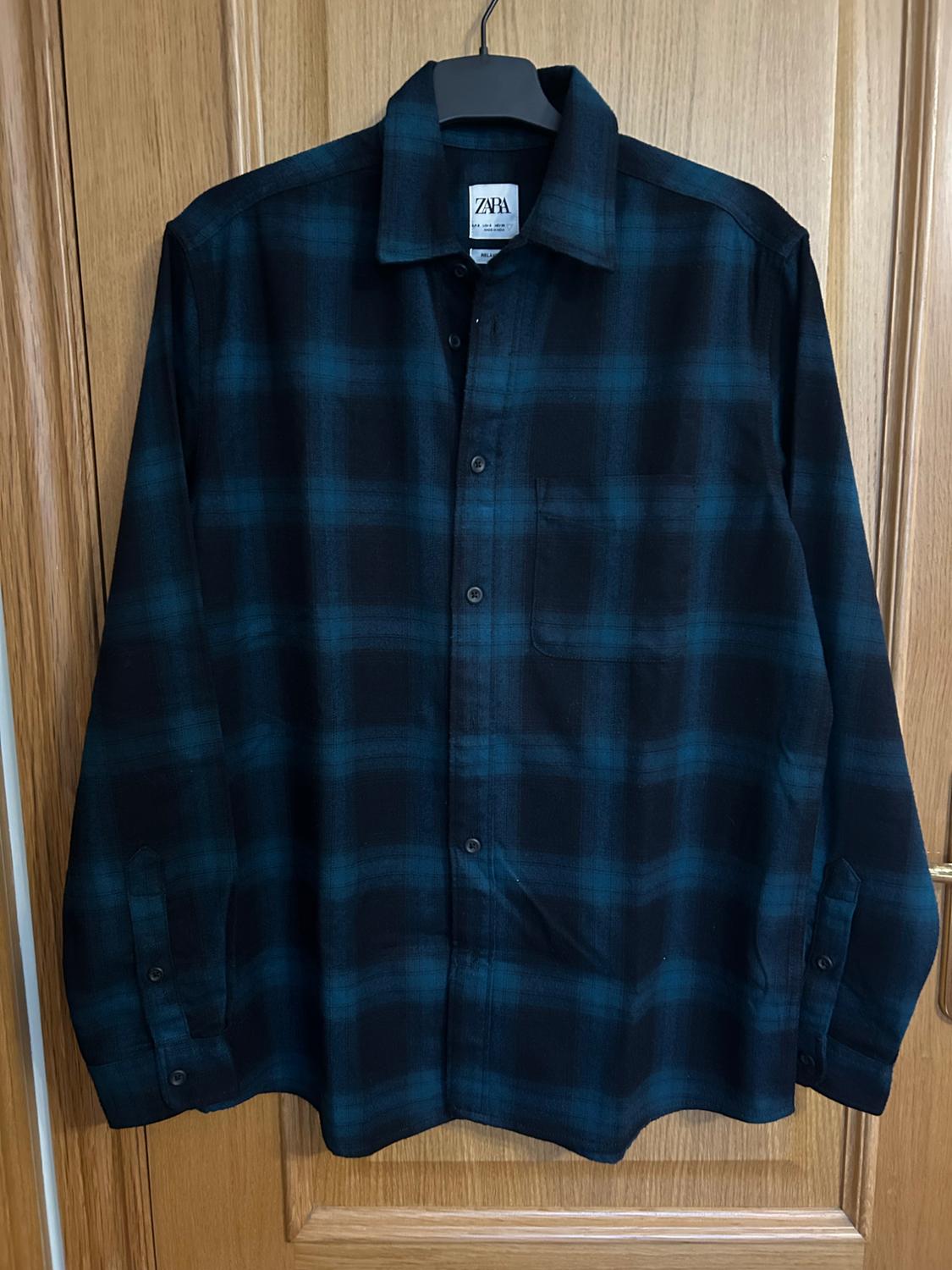 Blue and black checked long-sleeved shirt. It's hanging on a hanger over a wooden door. The label reads ZARA.