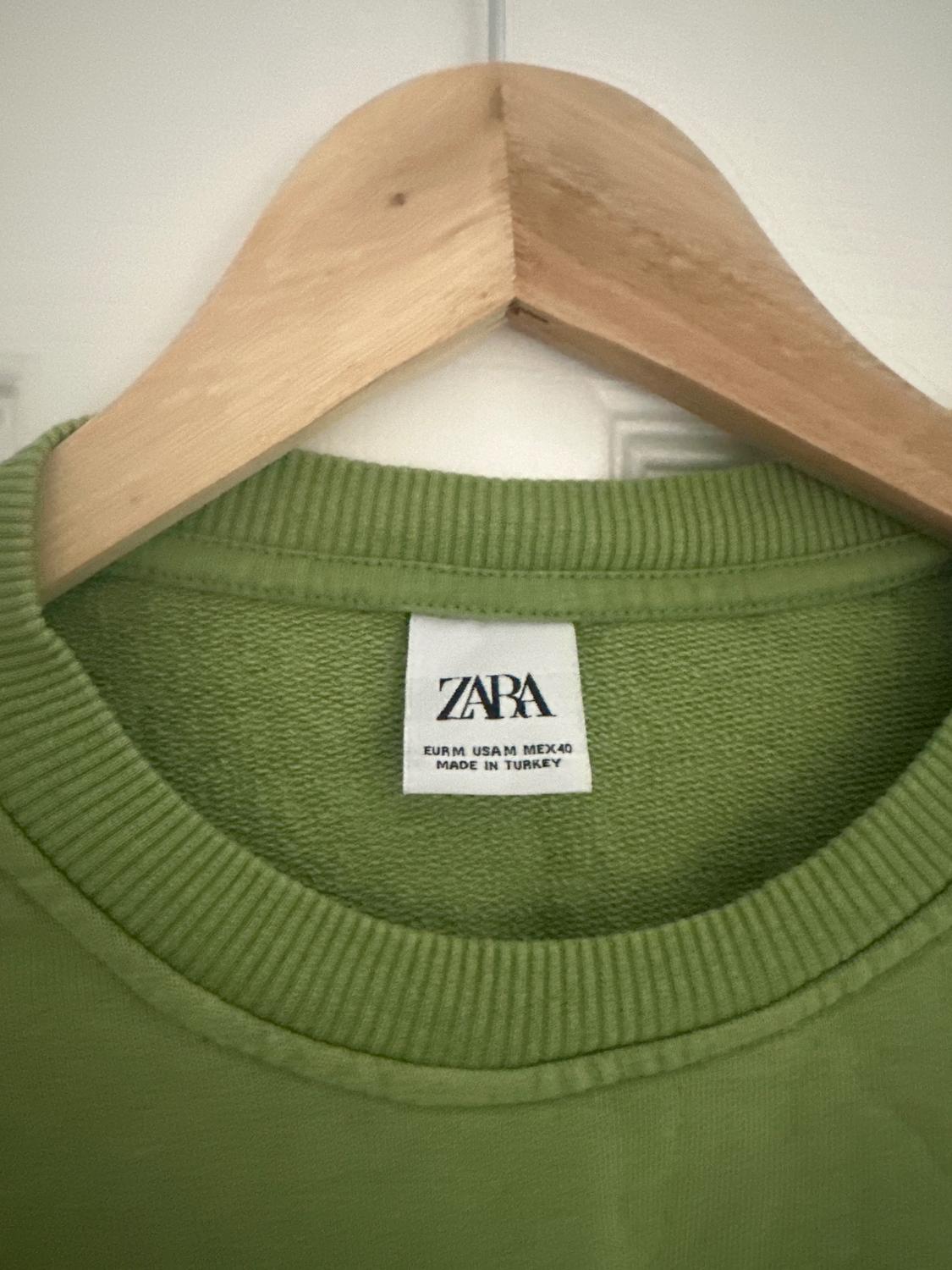 Label of a green t-shirt on a wooden hanger. Visible text: "ZARA EUR M USA M MEX 40 MADE IN TURKEY".