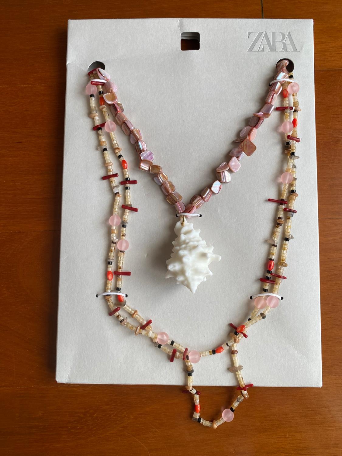 Pink beaded necklace with a white shell pendant on a card bearing the text ZARA, on a wooden table.