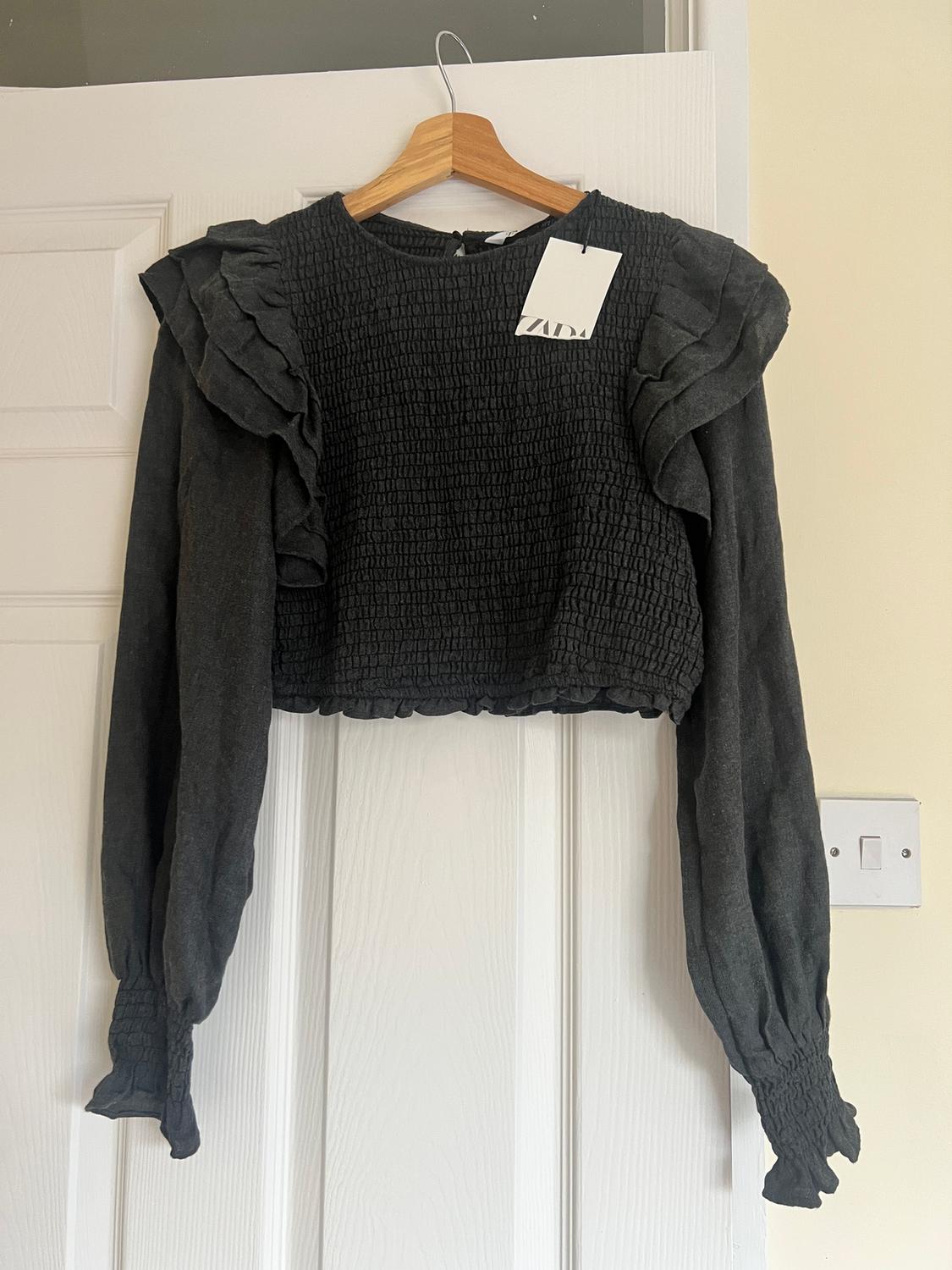 Black blouse with ruffled shoulders and long sleeves, hanging on a wooden hanger. Visible label with text "ZARA".
