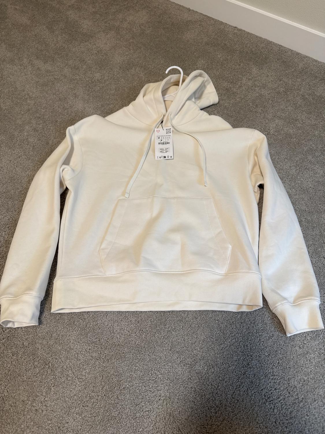 White hoodie with a front pocket, hanging on a hanger over a gray rug. Visible label with product details.