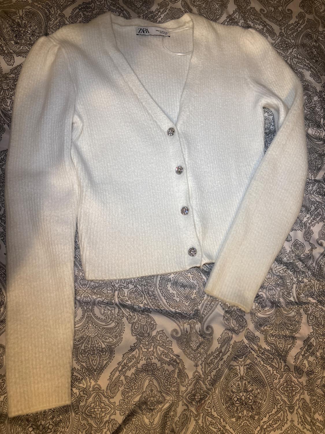 White knitted cardigan with sparkly buttons over grey-toned paisley print fabric. Visible label: ZARA.