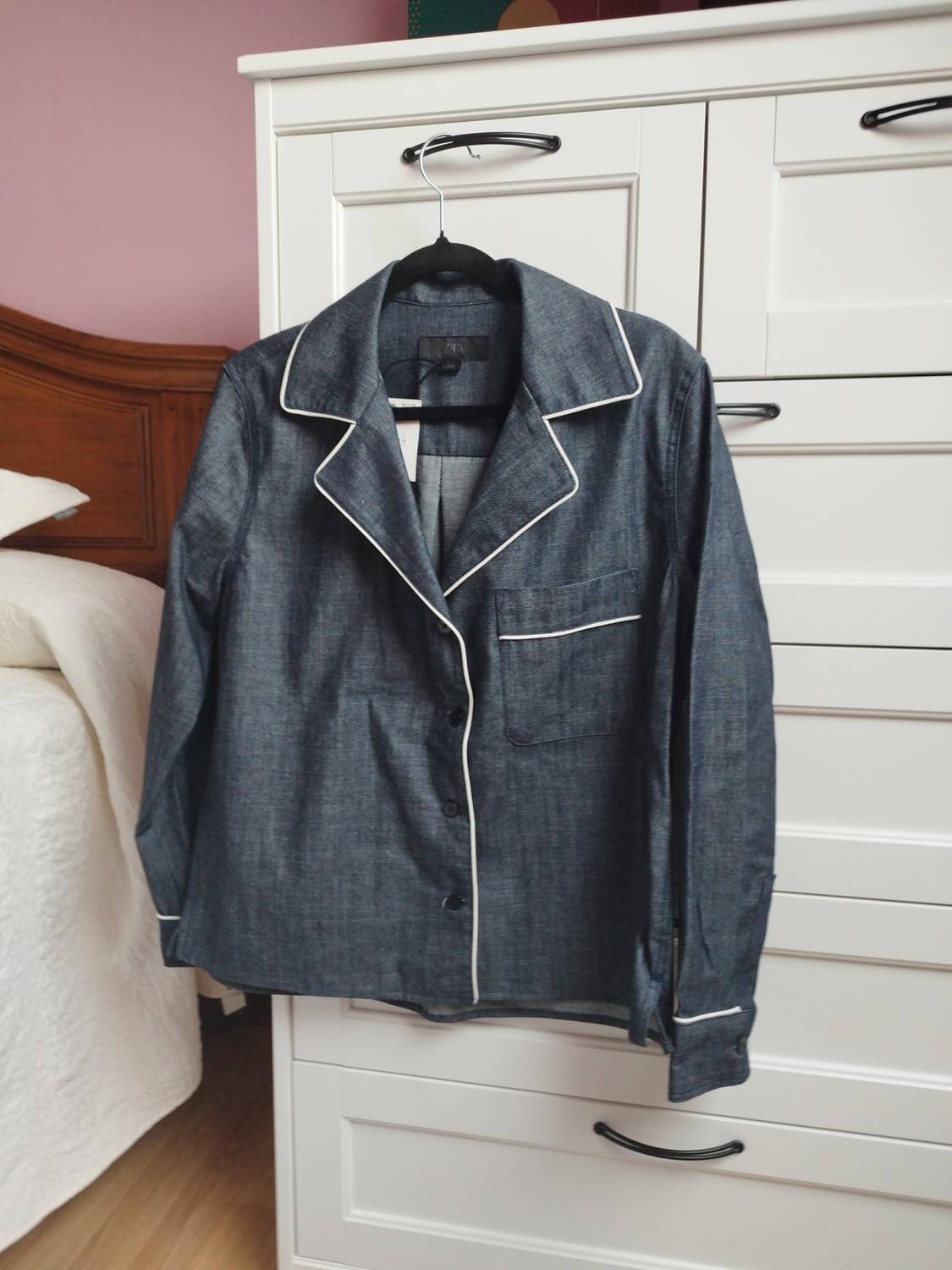 Blue pyjama shirt with white piping and a ZARA label, hanging on a white chest of drawers in a bedroom. Relaxed atmosphere.