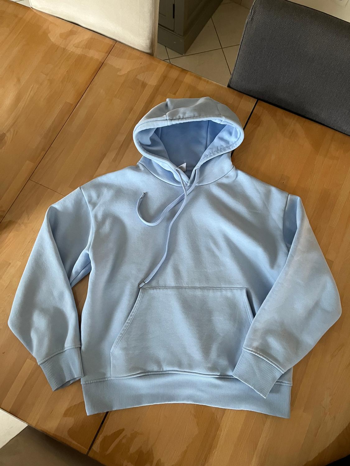 Light blue hoodie with a front pocket, placed on a light wooden table in an indoor setting.