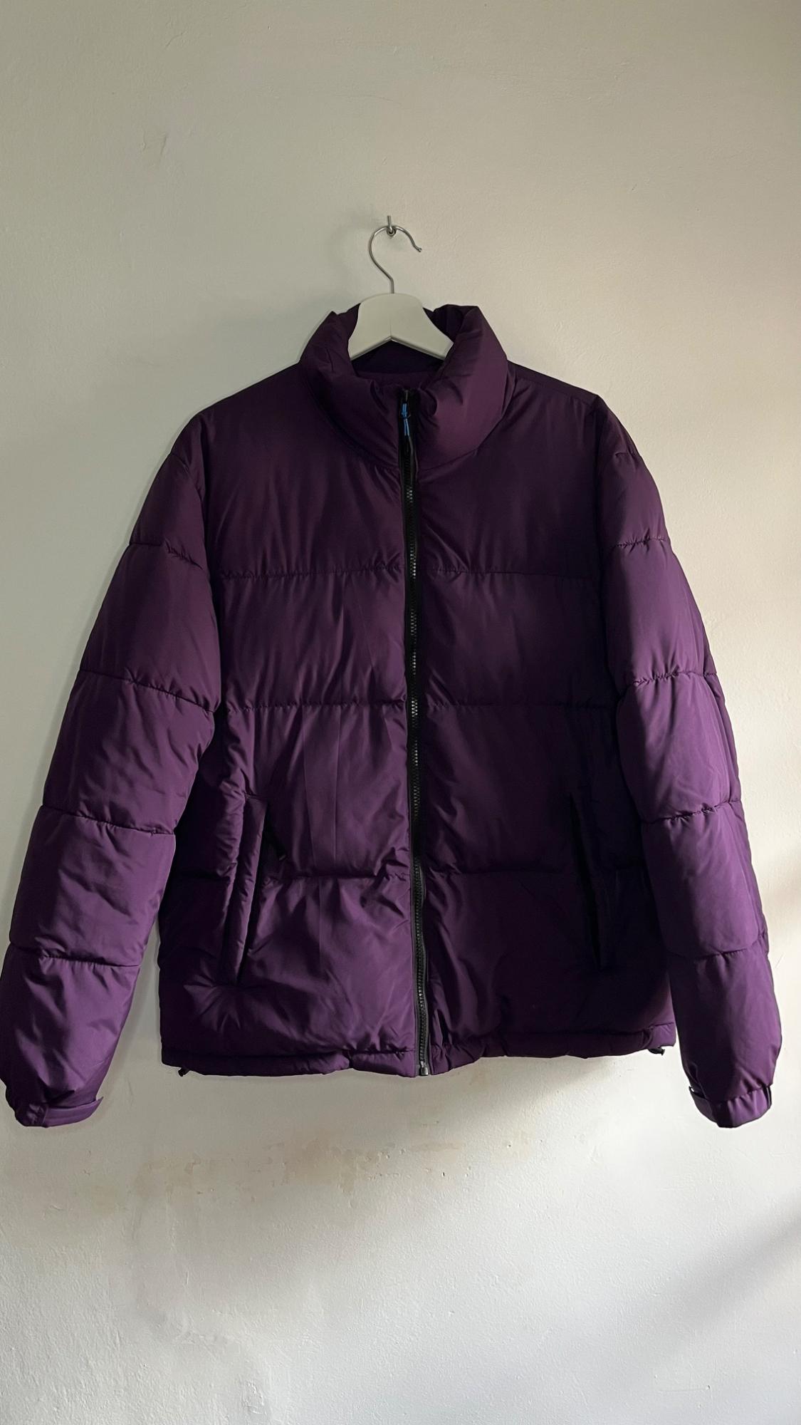 Purple padded jacket hanging on a white hanger against a light wall. Features a front zip and side pockets.