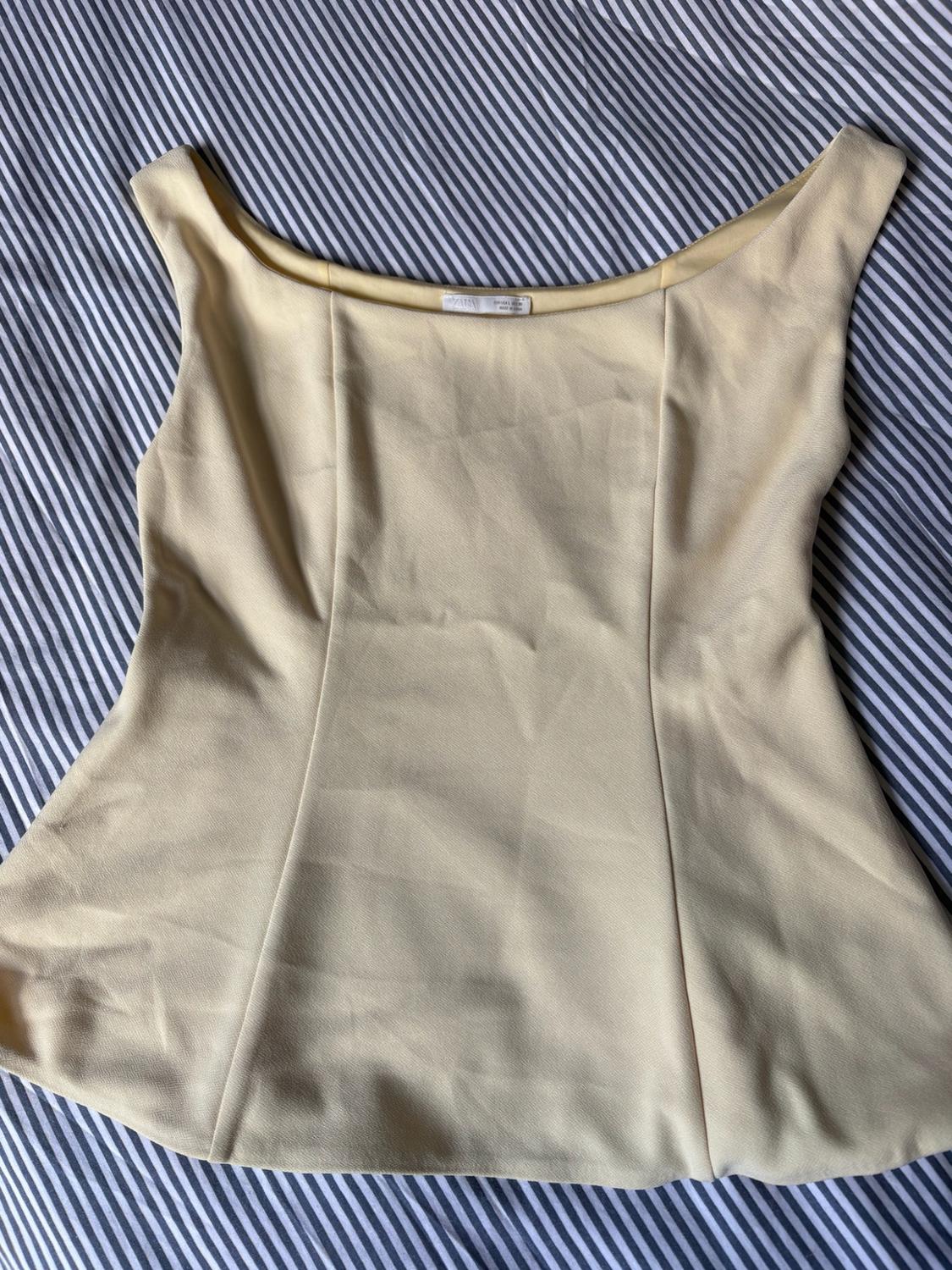 Light yellow sleeveless top with a wide neckline and vertical seams, laid out on a black and white striped fabric background.