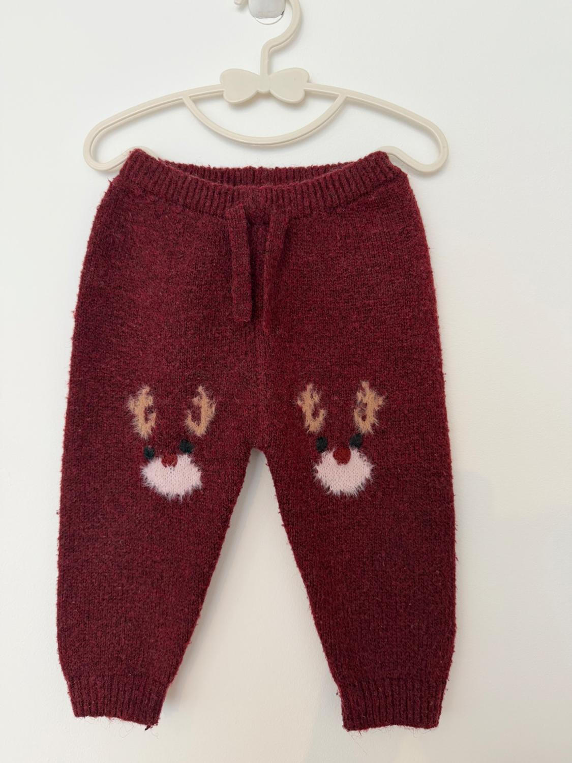 Maroon knitted children's trousers with reindeer faces, hanging on a white hanger with a bow against a white background. Cosy, winter style.