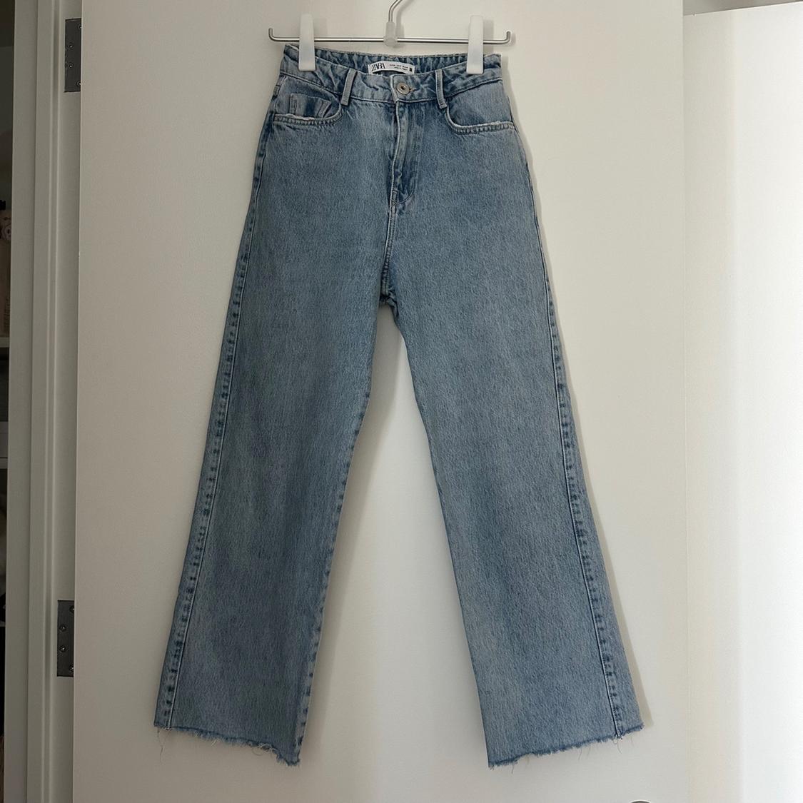 Light blue straight-cut jeans with a frayed hem, hanging on a hanger on a white door. ZARA label visible. Casual style.