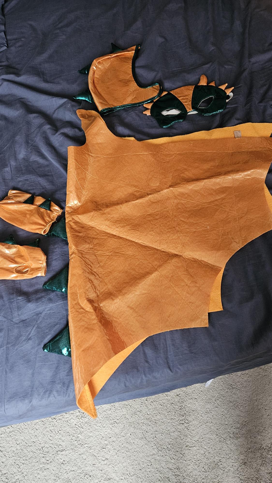 Orange and green dragon costume with wings, hood, and gloves, spread out on a bed with a gray comforter.