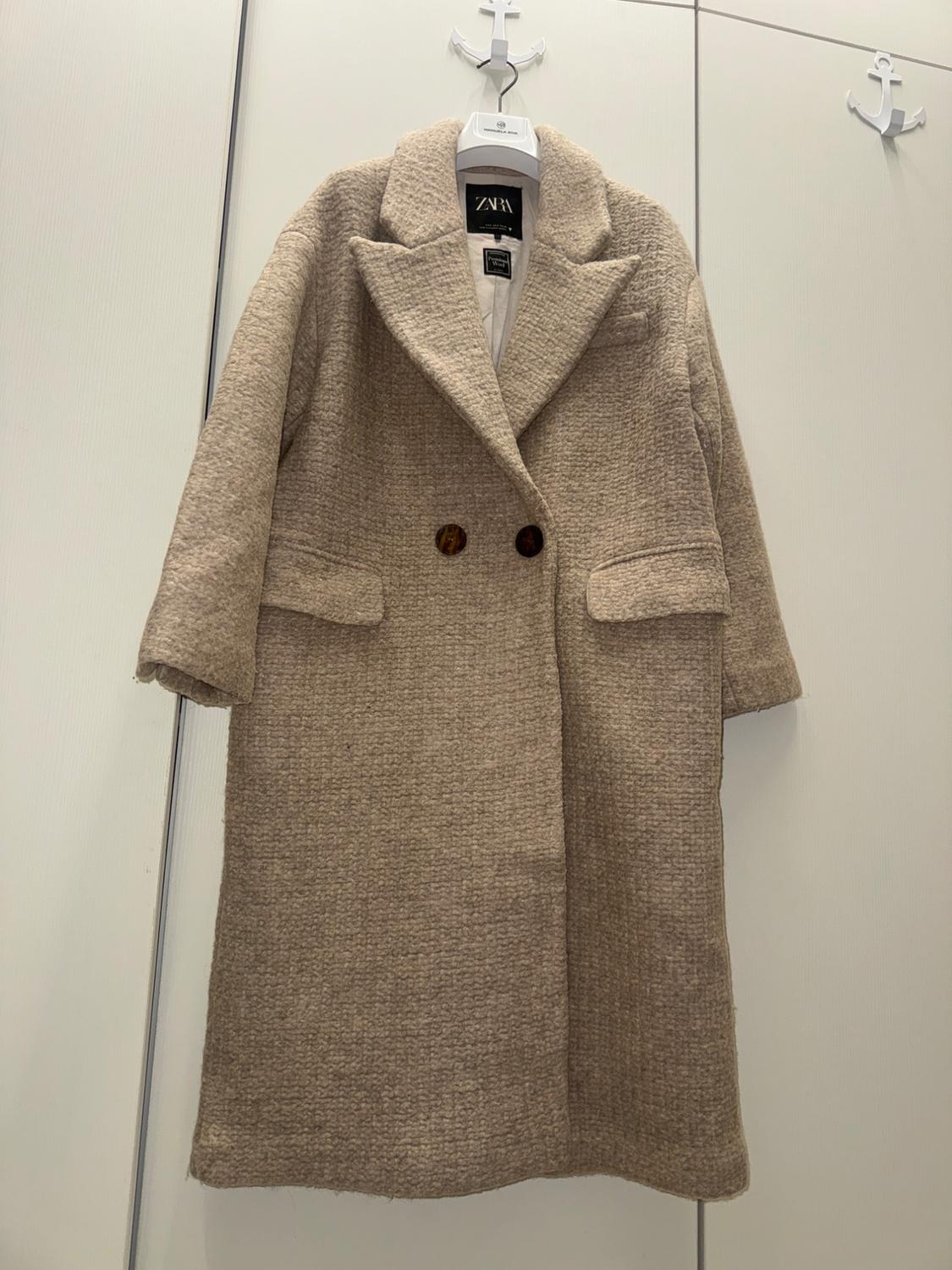 Beige wool coat hanging on a white hanger with an anchor. Visible Zara label. White wall background.