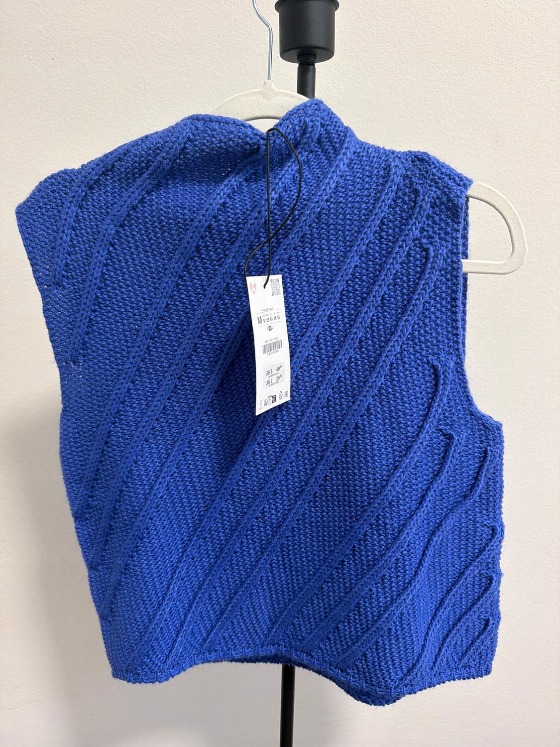Blue knitted vest with a diagonal line design, hanging on a white hanger. Visible label with text and barcode.
