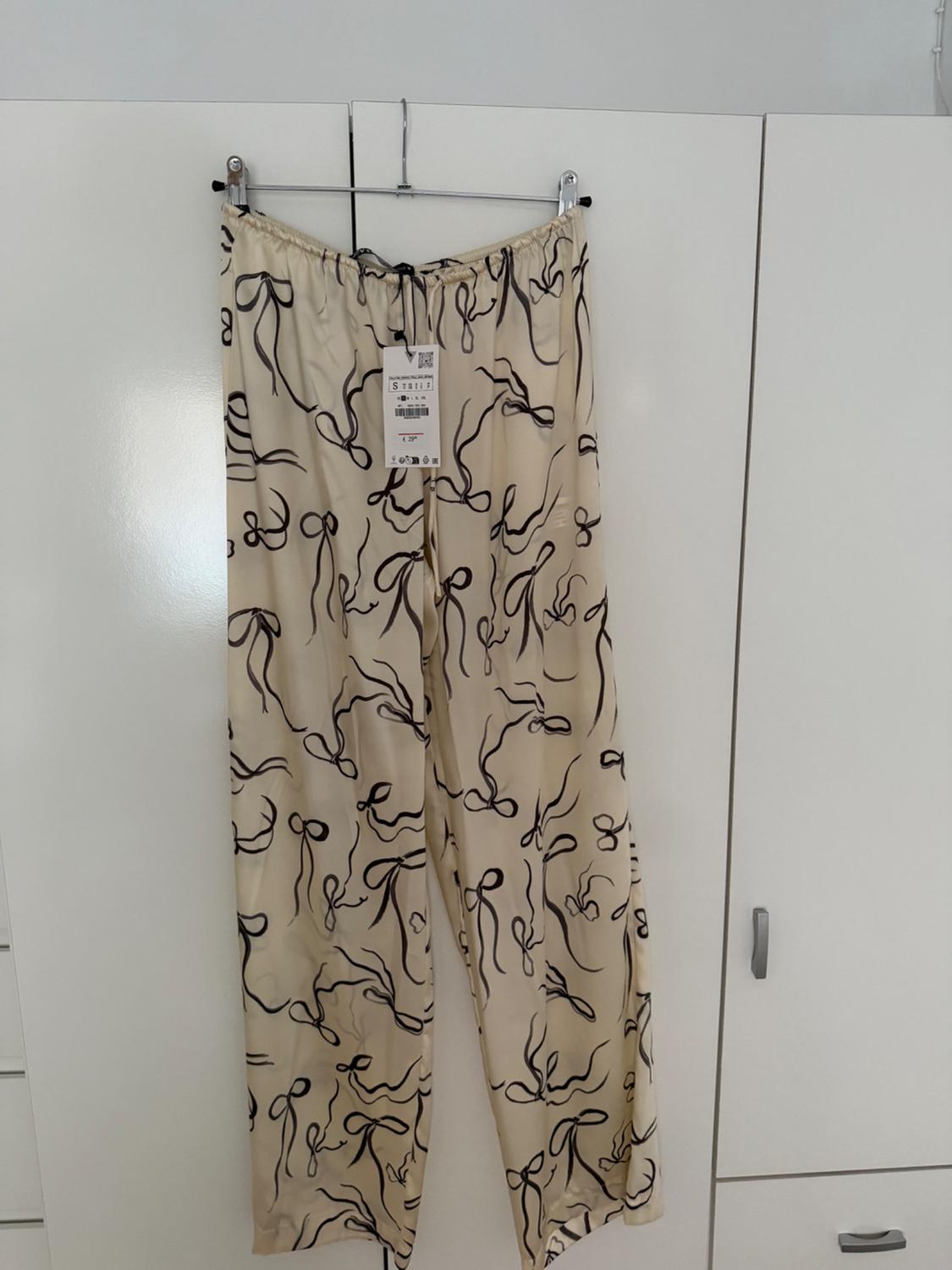 Beige trousers with a black bow print hanging on a hanger in front of a white wardrobe. Visible label on the waistband.